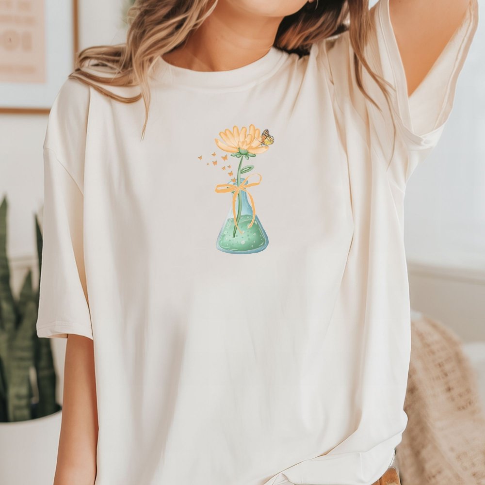 Ivory tee featuring a cute science-themed design with sunflower in a laboratory flask, tied with a bow, and a butterfly perched on top. Perfect blend of nature and science for a casual, fun look.