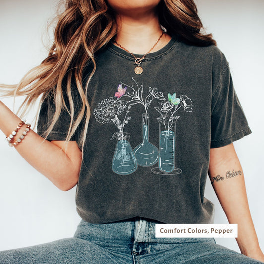 Comfort Colors tee in Pepper featuring a science-themed design with beakers, flasks, and flowers, accented by butterflies. A perfect combination of botanical and scientific elements in pastel colors on a trendy relaxed-fit shirt.