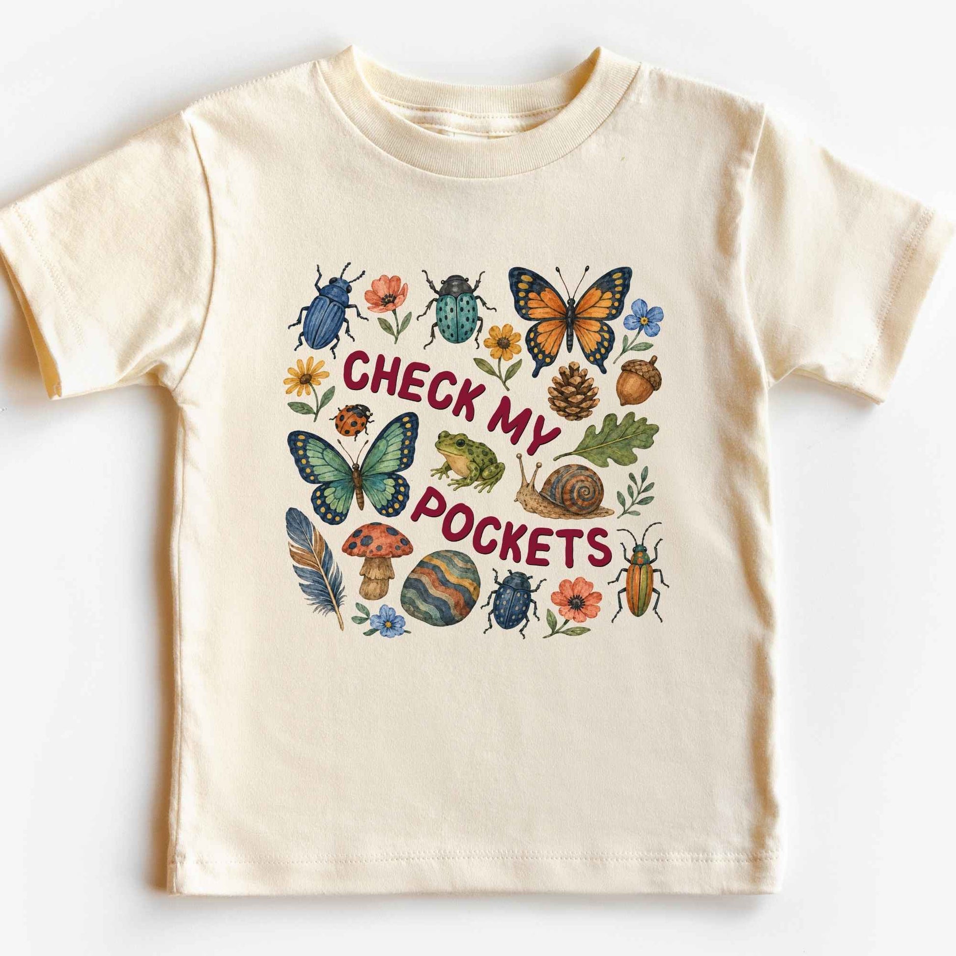 A cream-colored short-sleeve t-shirt with a colorful nature-themed graphic on the front. The graphic features various insects, flowers, a frog, a snail, pinecones, and mushrooms arranged around the red text 'CHECK MY POCKETS'.
