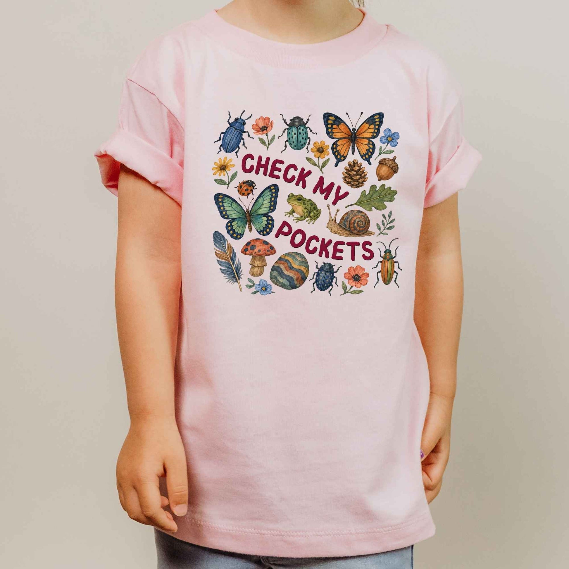 A child wearing a light pink short-sleeve t-shirt with a colorful graphic on the front that reads 'CHECK MY POCKETS' surrounded by illustrated insects, flowers, acorns, and mushrooms, photographed against a plain light gray background.