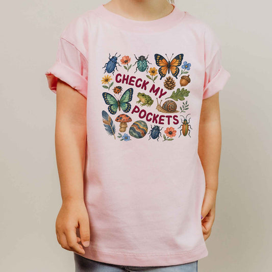 A child wearing a light pink short-sleeve t-shirt with a colorful graphic on the front that reads 'CHECK MY POCKETS' surrounded by illustrated insects, flowers, acorns, and mushrooms, photographed against a plain light gray background.