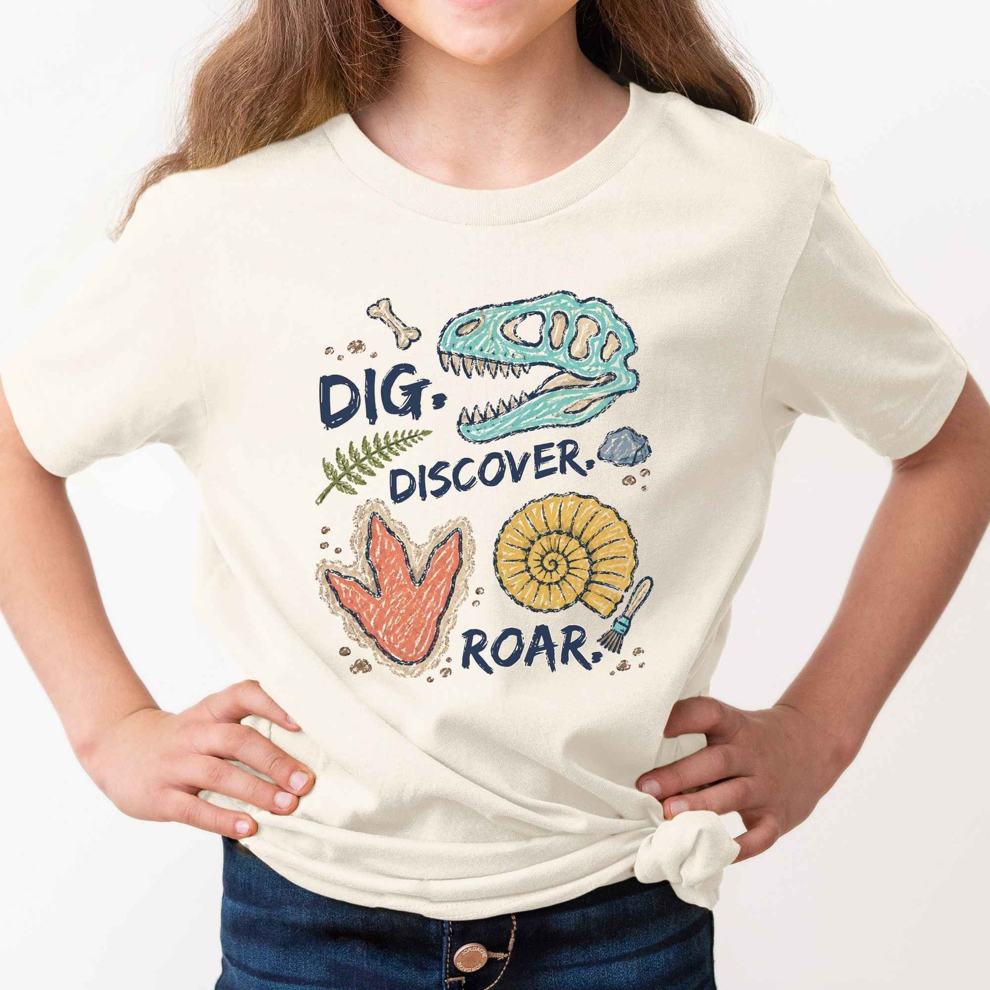 A child wearing a cream-colored t-shirt with a colorful dinosaur fossil graphic and the words 'DIG. DISCOVER.