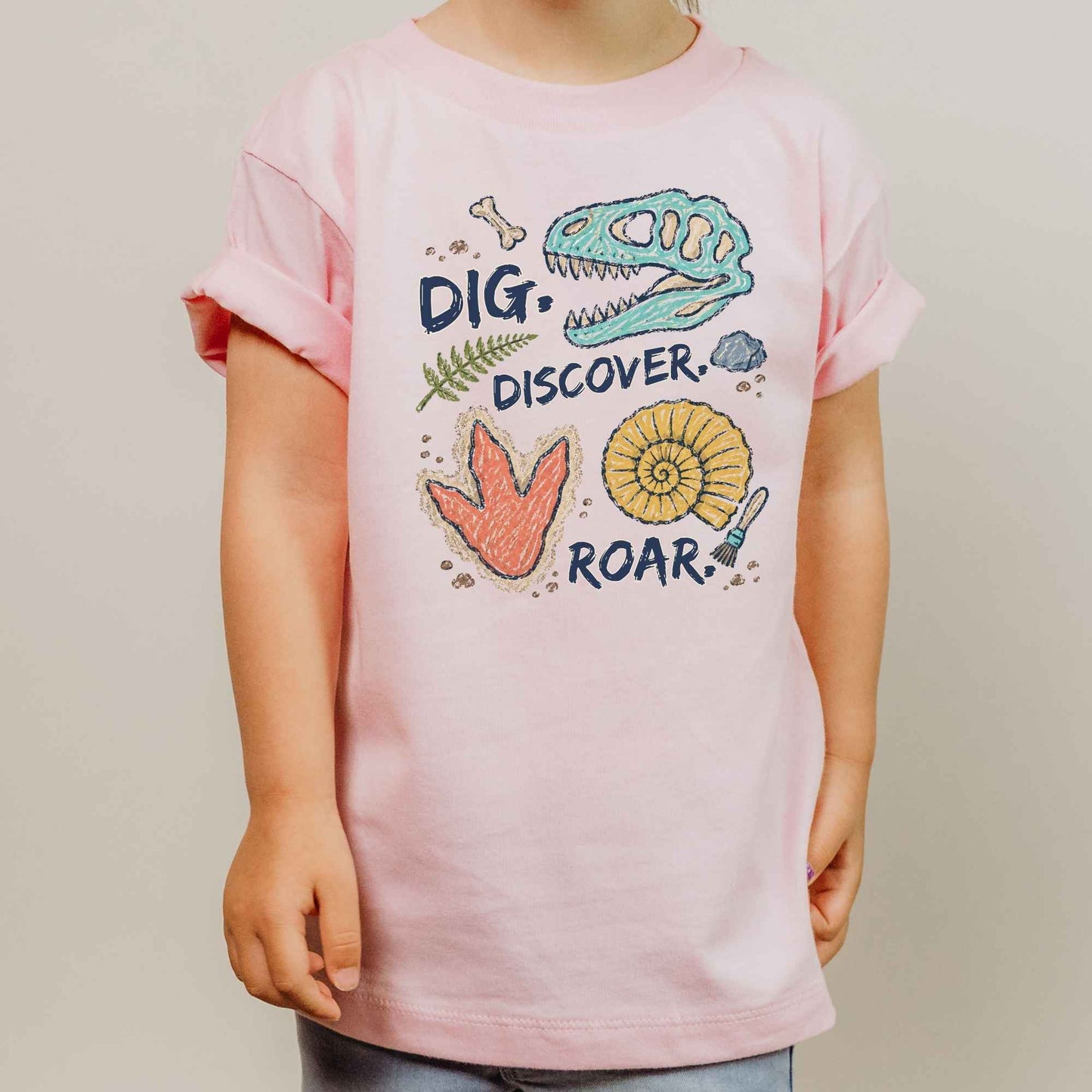 A child wearing a light pink short-sleeve t-shirt with a colorful dinosaur-themed graphic on the front that reads 'DIG. DISCOVER.