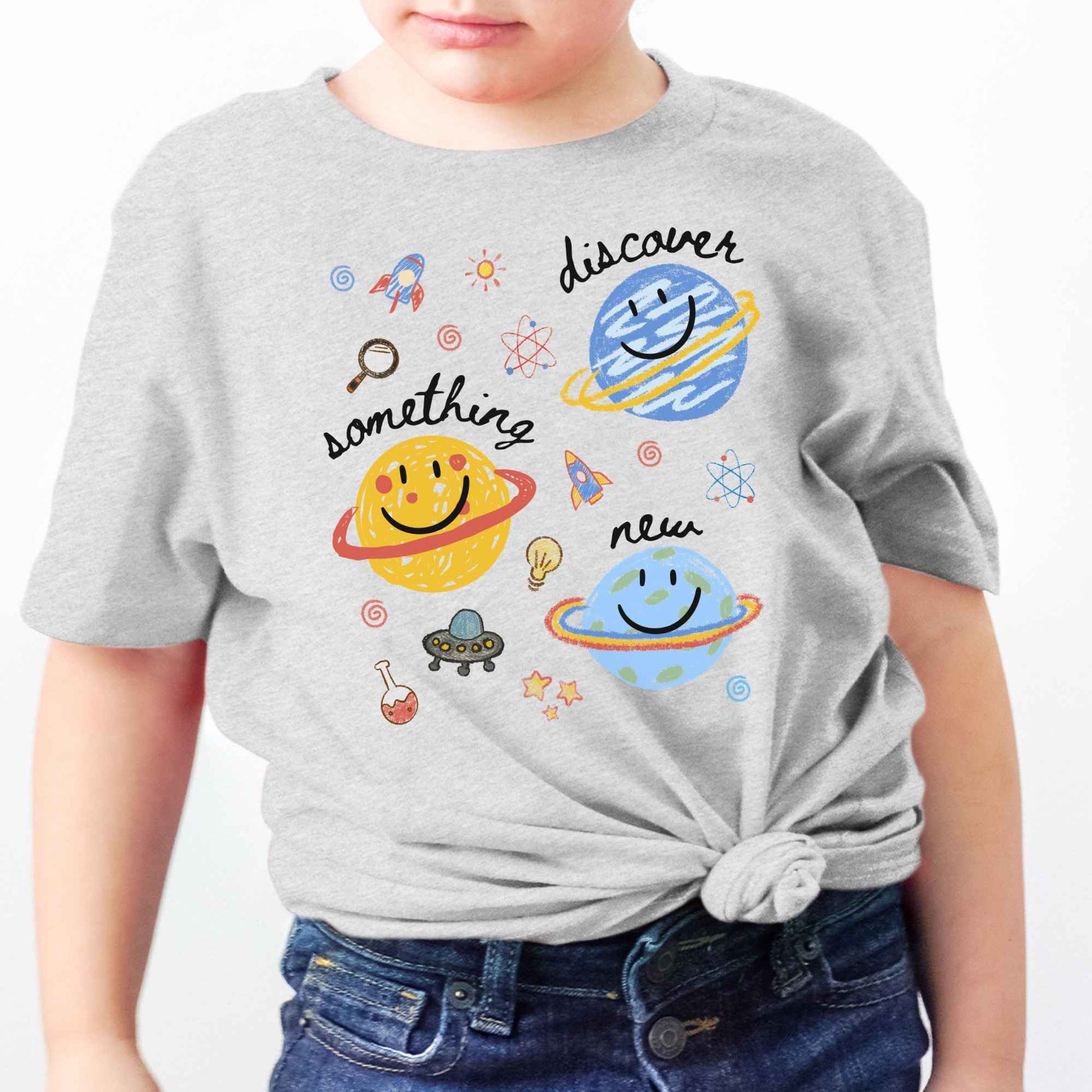 A child wearing a light heather gray t-shirt with a colorful space-themed graphic print featuring planets, rockets, stars, and the words 'discover something new'.