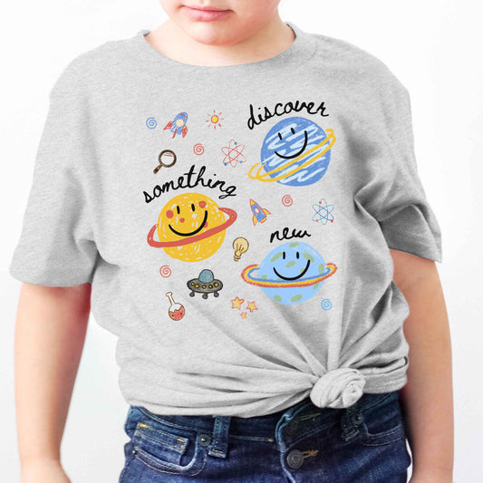A child wearing a light heather gray t-shirt with a colorful space-themed graphic print featuring planets, rockets, stars, and the words 'discover something new'.