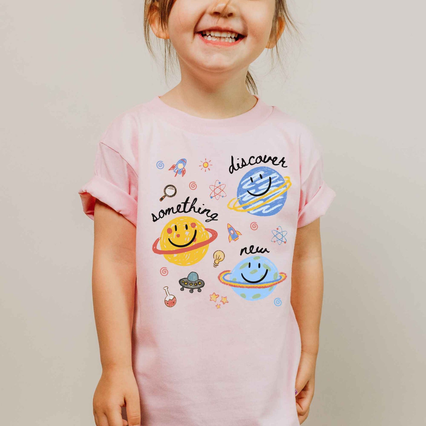 A young child wearing a light pink short-sleeve t-shirt with a colorful space-themed graphic print featuring planets, rockets, stars, and the words 'discover something new'.