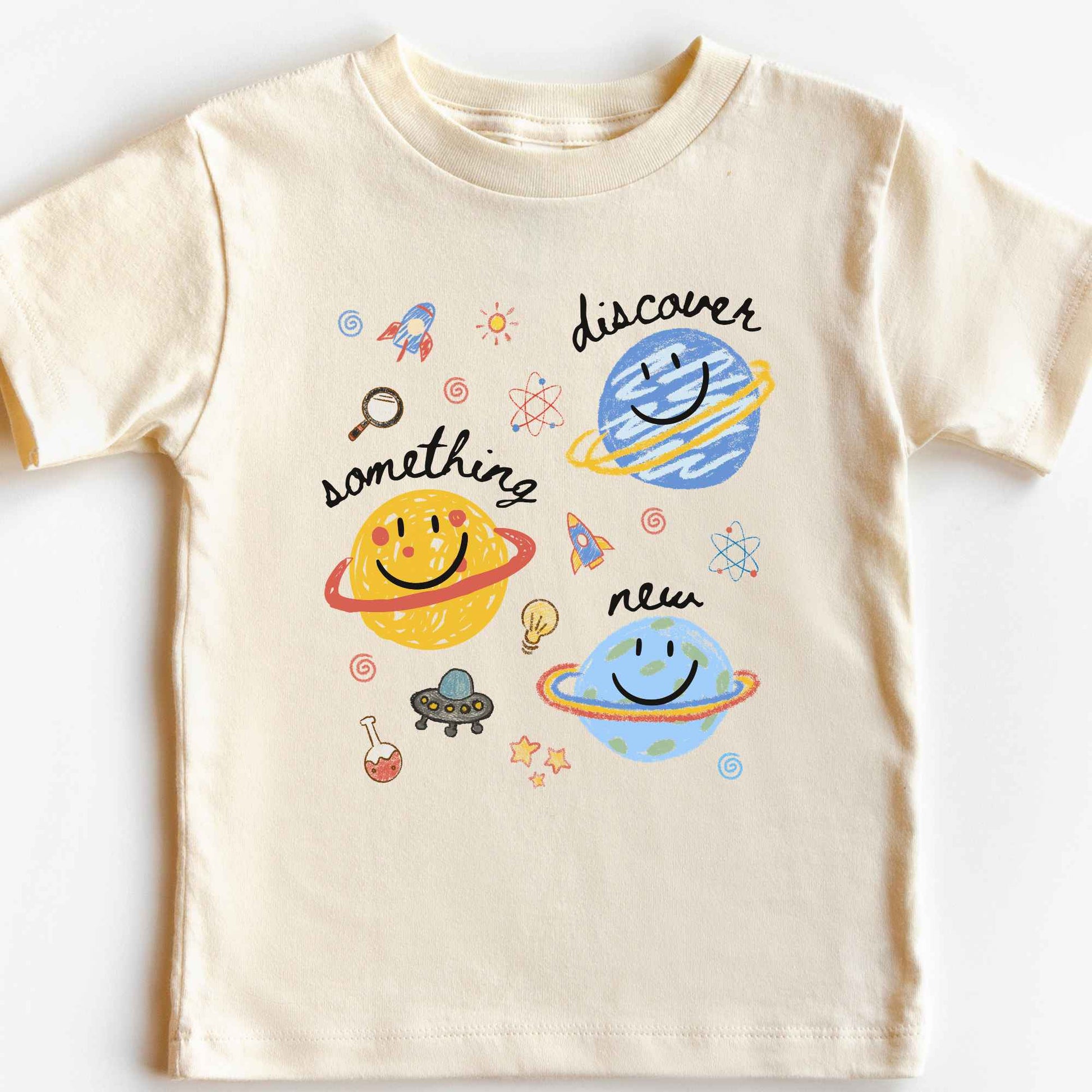 A cream-colored short-sleeve baby t-shirt featuring a colorful cartoon space graphic with planets, rockets, and the words 'discover something new'.
