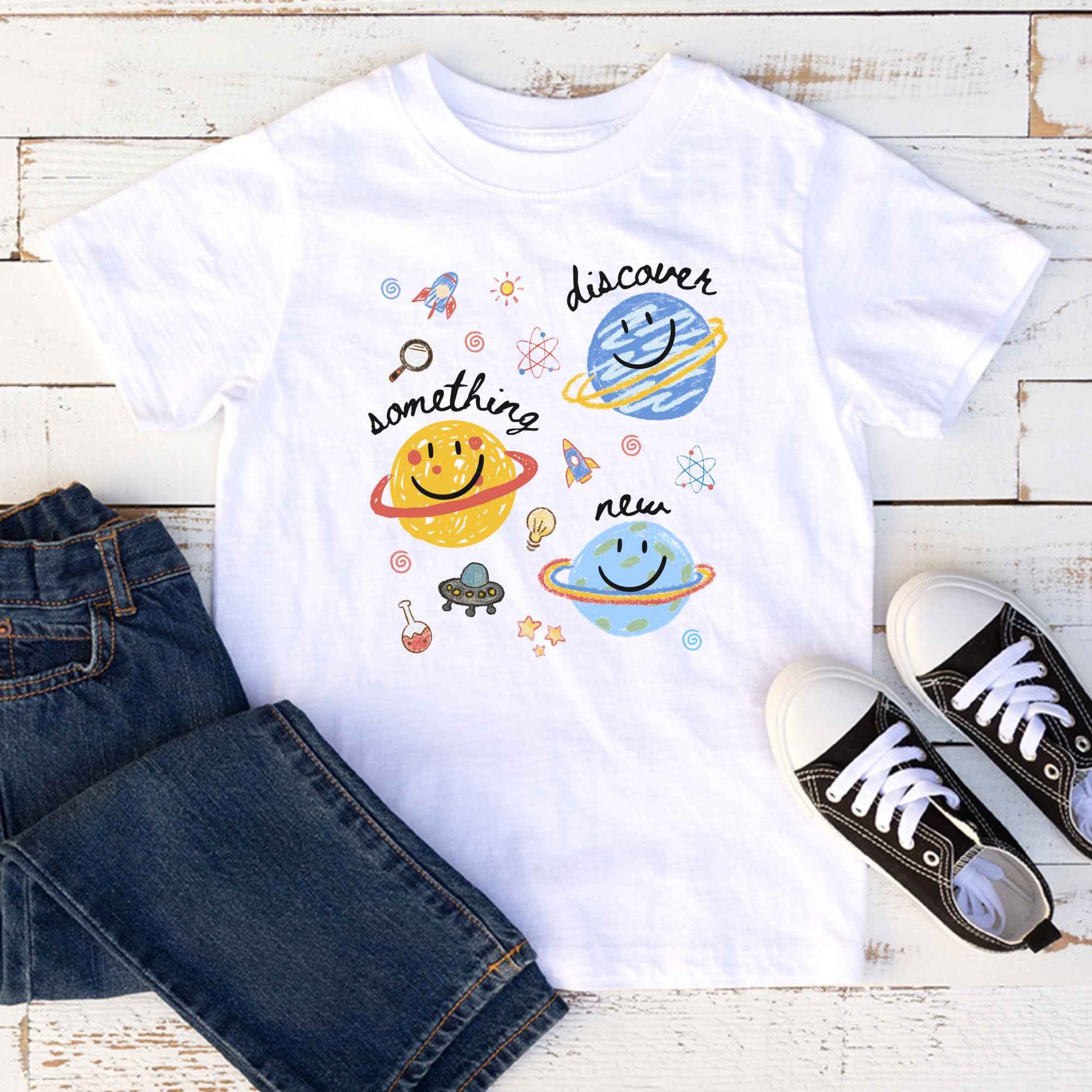 A white kids' t-shirt with a colorful space-themed graphic print featuring planets, rockets, and the words 'discover something new', styled in a flat lay with blue jeans and black sneakers on a white wood background.