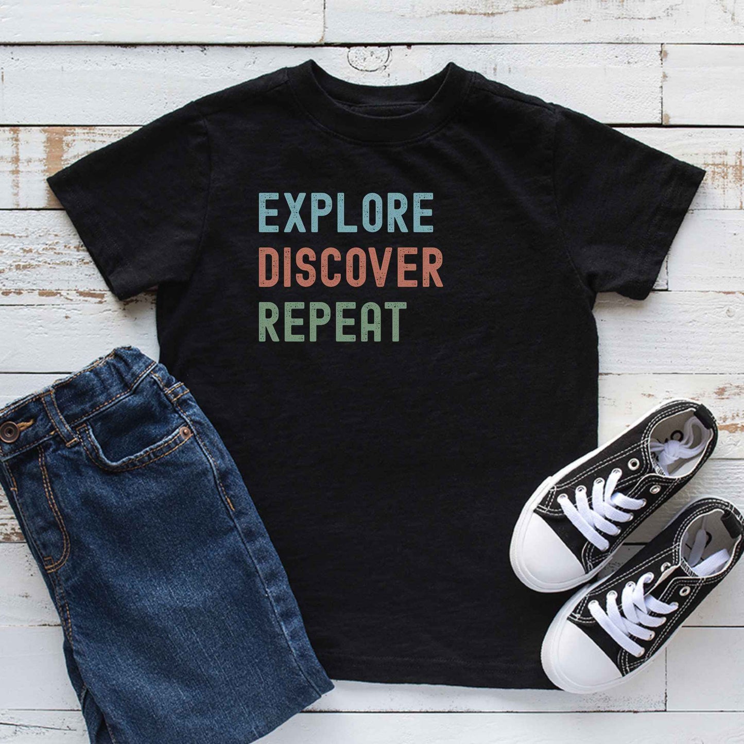 A black graphic T-shirt laid flat on a white wood plank background, styled with blue jeans and black-and-white sneakers. The shirt has the words 'EXPLORE DISCOVER REPEAT' printed on the front in light blue, muted orange, and sage green.