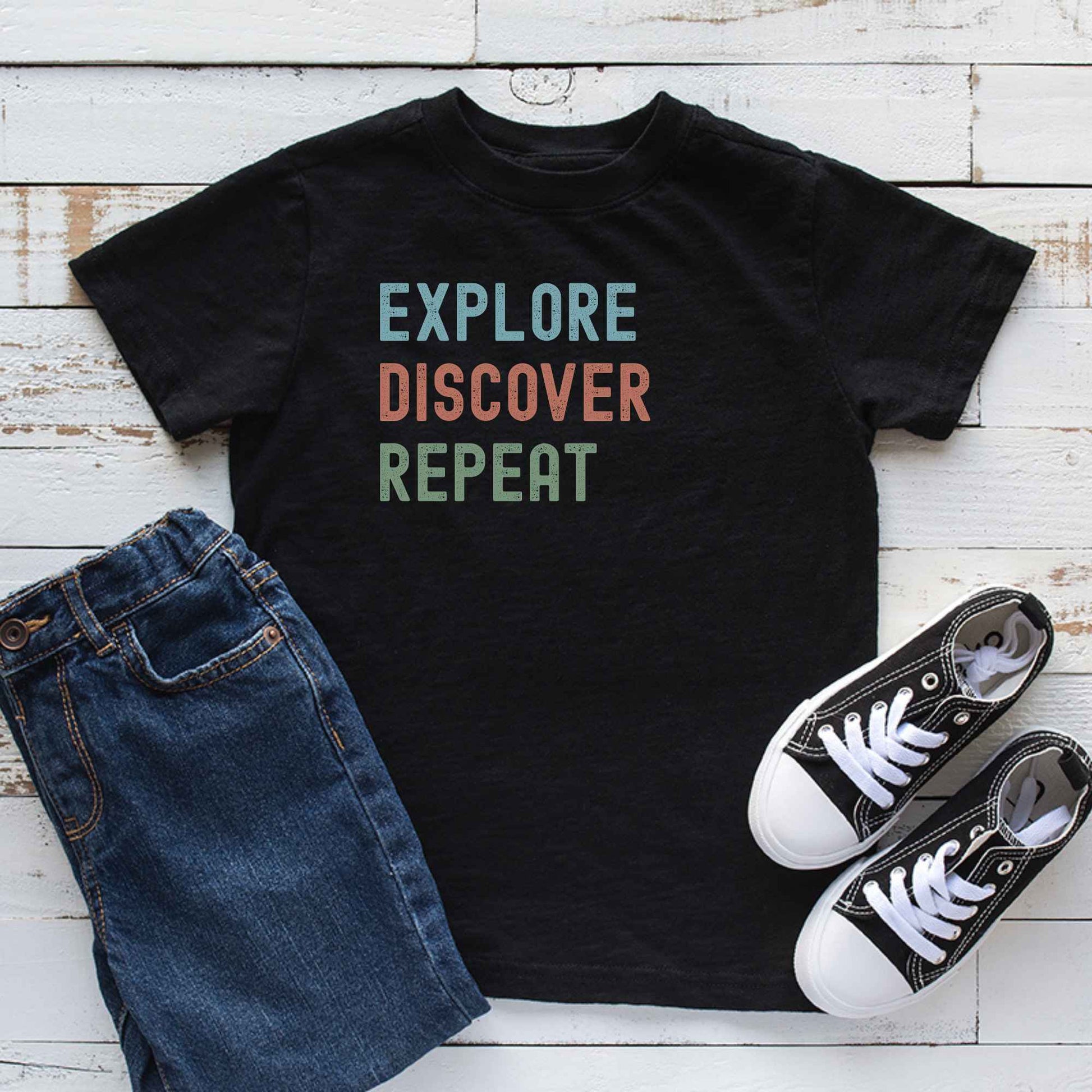 A black graphic T-shirt laid flat on a white wood plank background, styled with blue jeans and black-and-white sneakers. The shirt has the words 'EXPLORE DISCOVER REPEAT' printed on the front in light blue, muted orange, and sage green.
