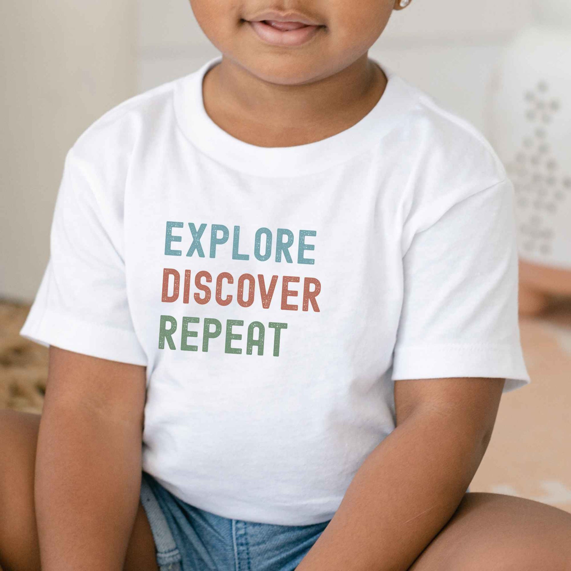 A child wearing a white short-sleeve T-shirt with the words 'EXPLORE DISCOVER REPEAT' printed in muted blue, orange, and green across the chest, paired with denim shorts in a softly lit indoor setting.