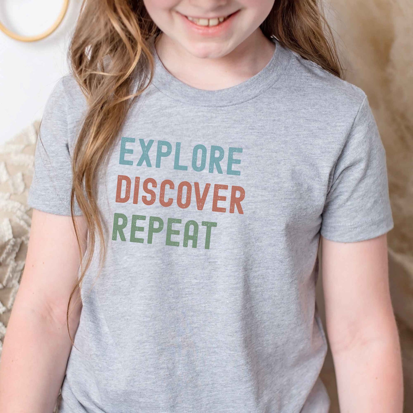 A child wearing a heather gray short-sleeve t-shirt with the motivational text 'EXPLORE DISCOVER REPEAT' printed in teal, dusty orange, and sage green block letters across the chest.