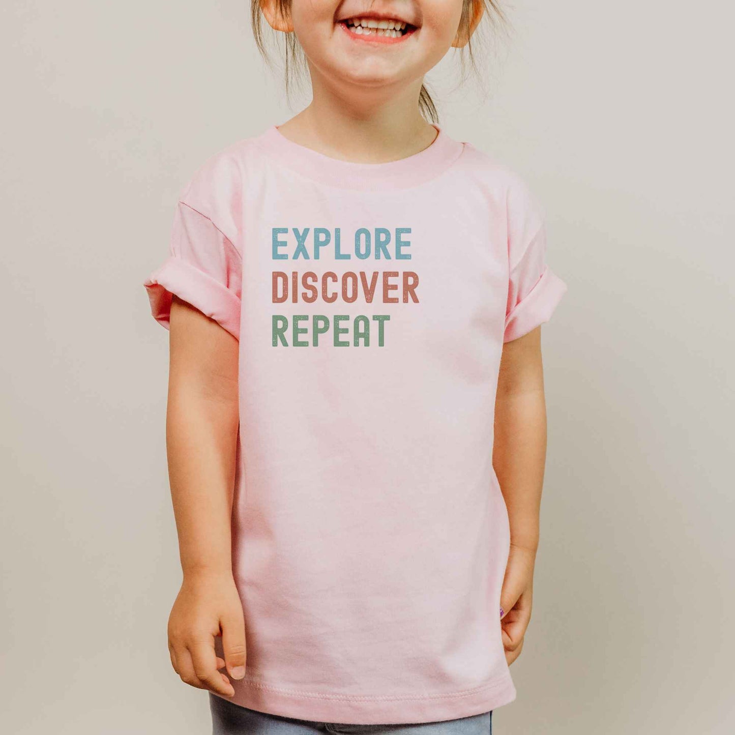 A young child wearing a light pink t-shirt with the text 'EXPLORE DISCOVER REPEAT' printed on the chest in blue, orange, and green lettering, set against a plain light gray background.
