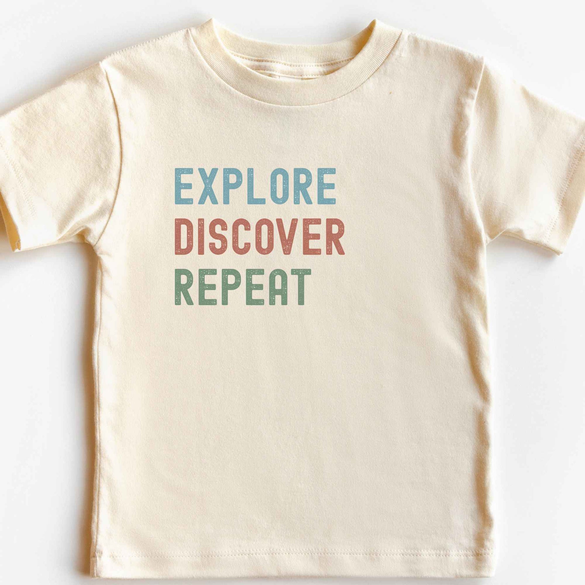 A cream-colored short-sleeve t-shirt with the motivational phrase 'EXPLORE DISCOVER REPEAT' printed on the chest in blue, muted red, and sage green block letters, laid flat against a white background.