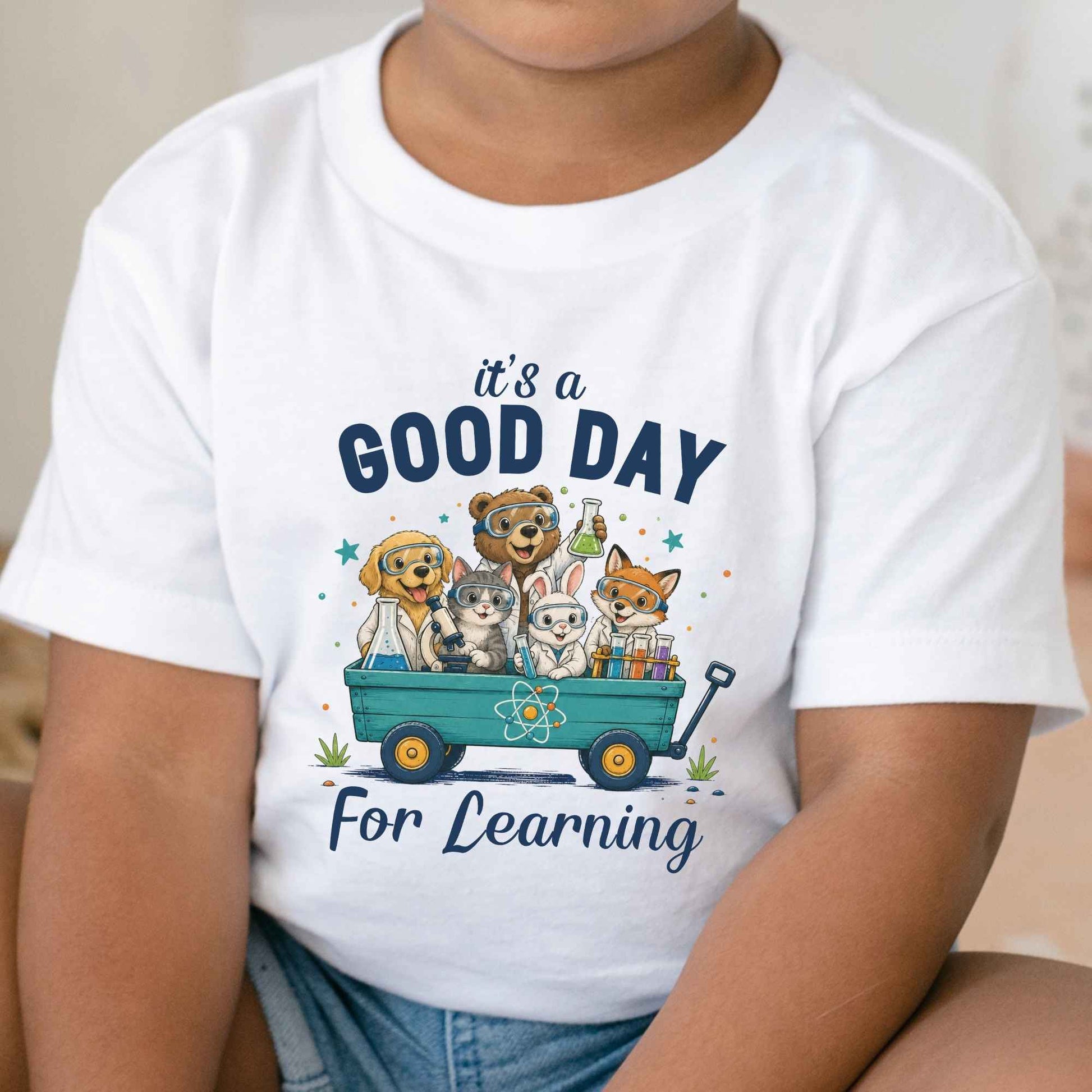A child wearing a white short-sleeve T-shirt with a colorful cartoon graphic of animals in a wagon and the text 'it's a GOOD DAY For Learning'.