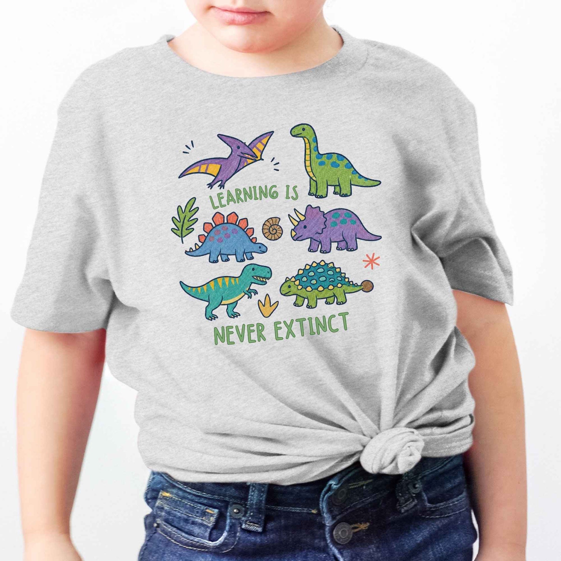 A child wearing a light heather gray t-shirt with a colorful dinosaur graphic and the text 'LEARNING IS NEVER EXTINCT'. The child is also wearing blue jeans, shown from the waist up against a plain white background.