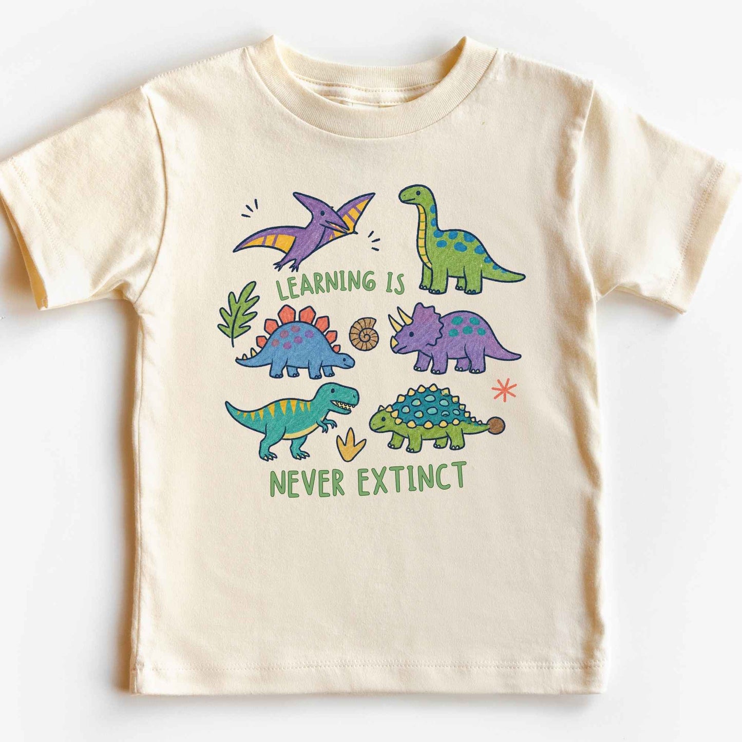 A cream-colored short-sleeve t-shirt with a colorful cartoon dinosaur graphic and the text 'LEARNING IS NEVER EXTINCT' printed on the front, displayed flat against a white background.