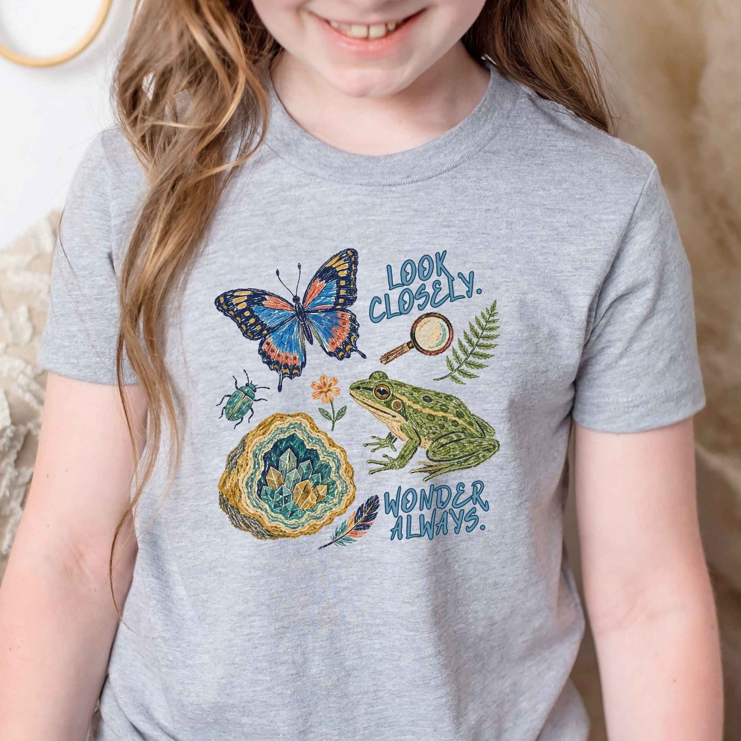 A child wearing a heather gray graphic T-shirt featuring a colorful nature print with a butterfly, frog, geode, magnifying glass, fern, and the words 'LOOK CLOSER. WONDERS ALWAYS.