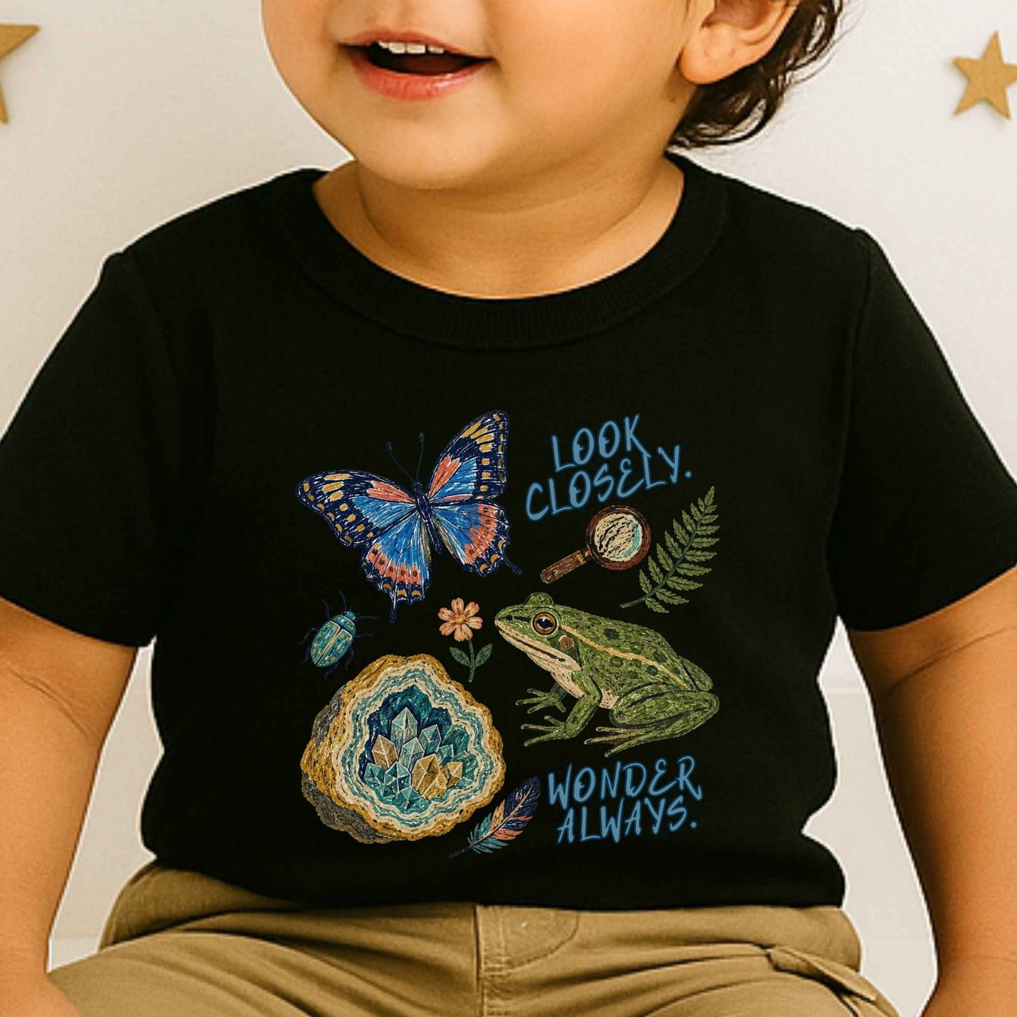 A toddler wearing a black short-sleeve graphic T-shirt with a colorful nature print featuring a butterfly, frog, geode, magnifying glass, fern, and the words 'LOOK CLOSELY. WONDER ALWAYS.