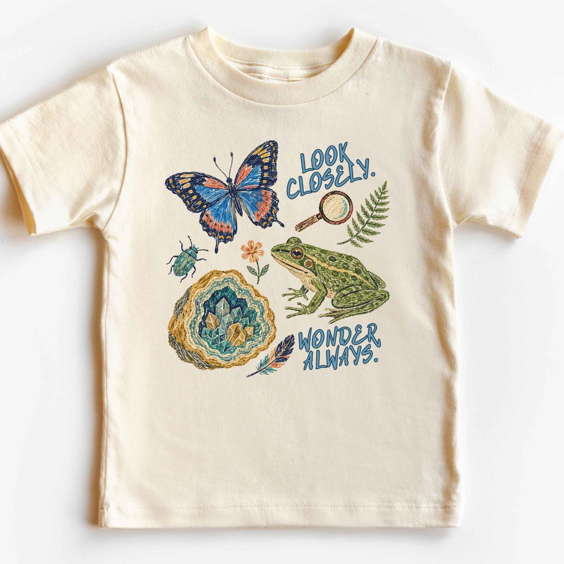 A cream-colored short-sleeve baby t-shirt with a colorful graphic print featuring a butterfly, frog, geode, beetle, magnifying glass, and fern. The text on the graphic reads 'LOOK CLOSELY.