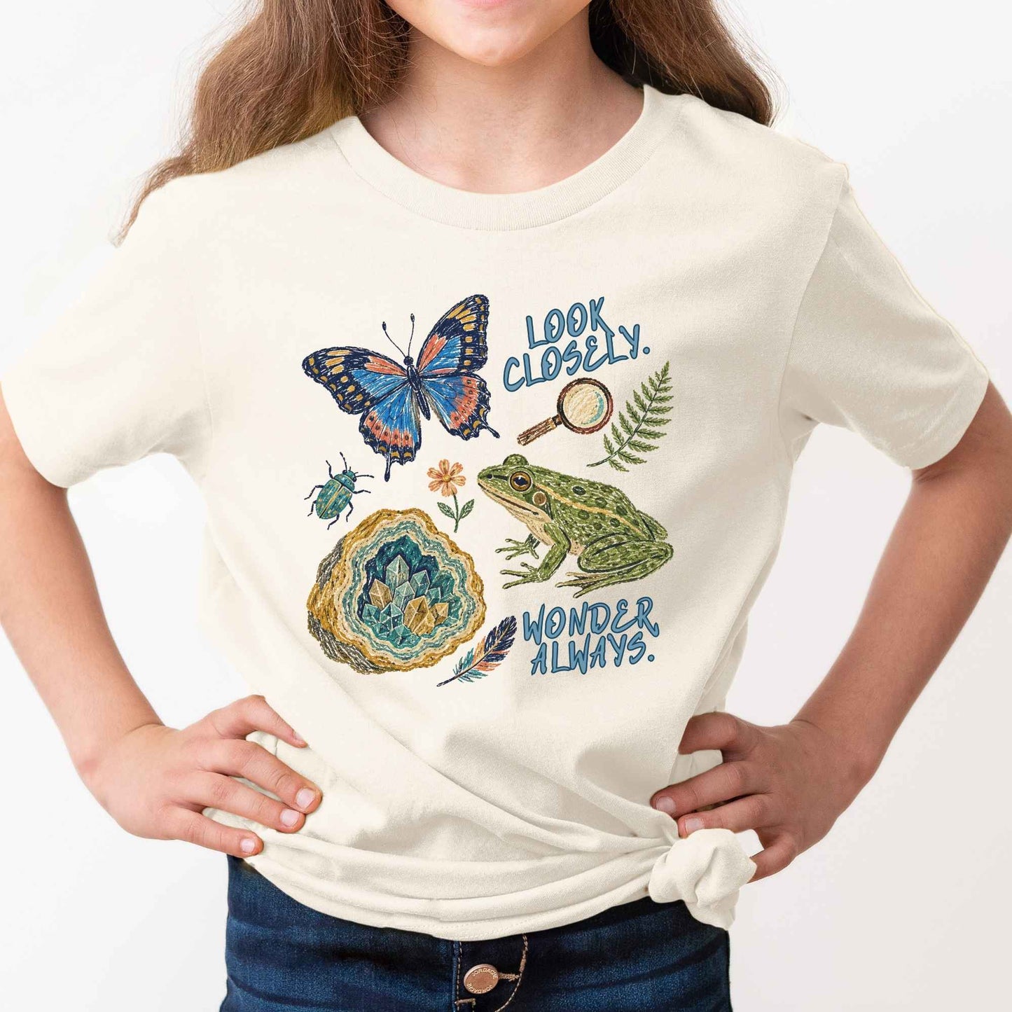 A child wearing a cream-colored short-sleeve t-shirt with a colorful nature-themed graphic print featuring a butterfly, frog, geode, magnifying glass, and the words 'LOOK CLOSETLY. WONDER ALWAYS.