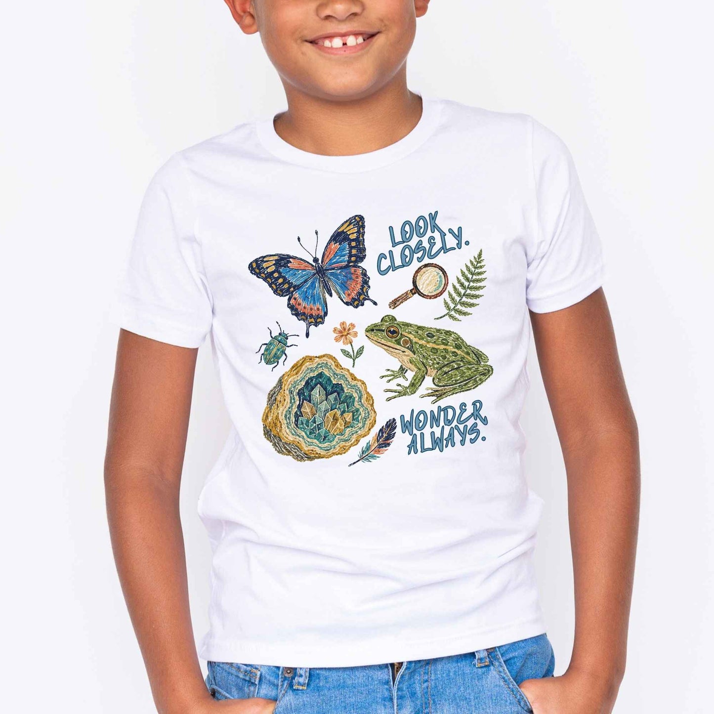 A child wearing a white short-sleeve graphic T-shirt with illustrations of a butterfly, frog, geode, fern, magnifying glass, and text reading 'LOOK CLOSELY. WONDER ALWAYS.