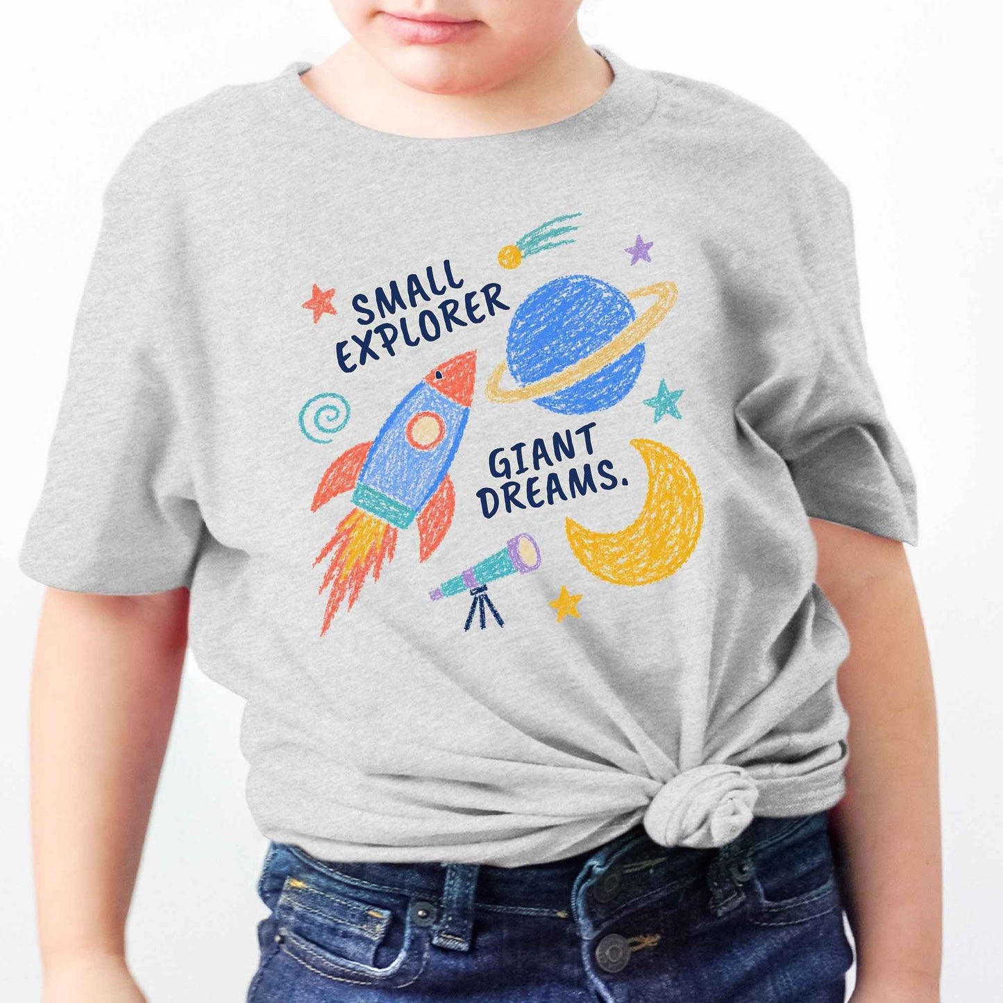 A child wearing a light heather gray t-shirt with a colorful space-themed graphic that reads 'SMALL EXPLORER GIANT DREAMS.' The shirt has a bright rocket, planet, moon, and telescope design, and is styled with a knot at the bottom.