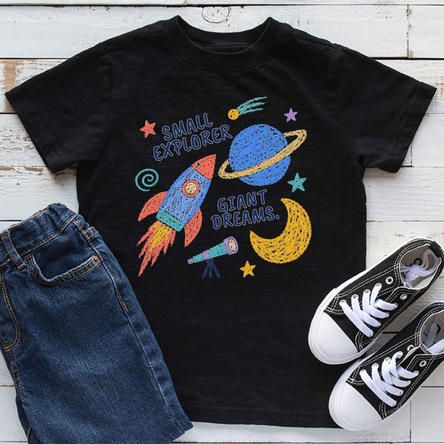 A black short-sleeve T-shirt laid flat on a white wood background, styled with blue jeans and black-and-white sneakers. The shirt features a colorful space-themed graphic with the words 'SMALL EXPLORER' and 'GIANT DREAMS,' showing a rocket, planet, moon, and stars.