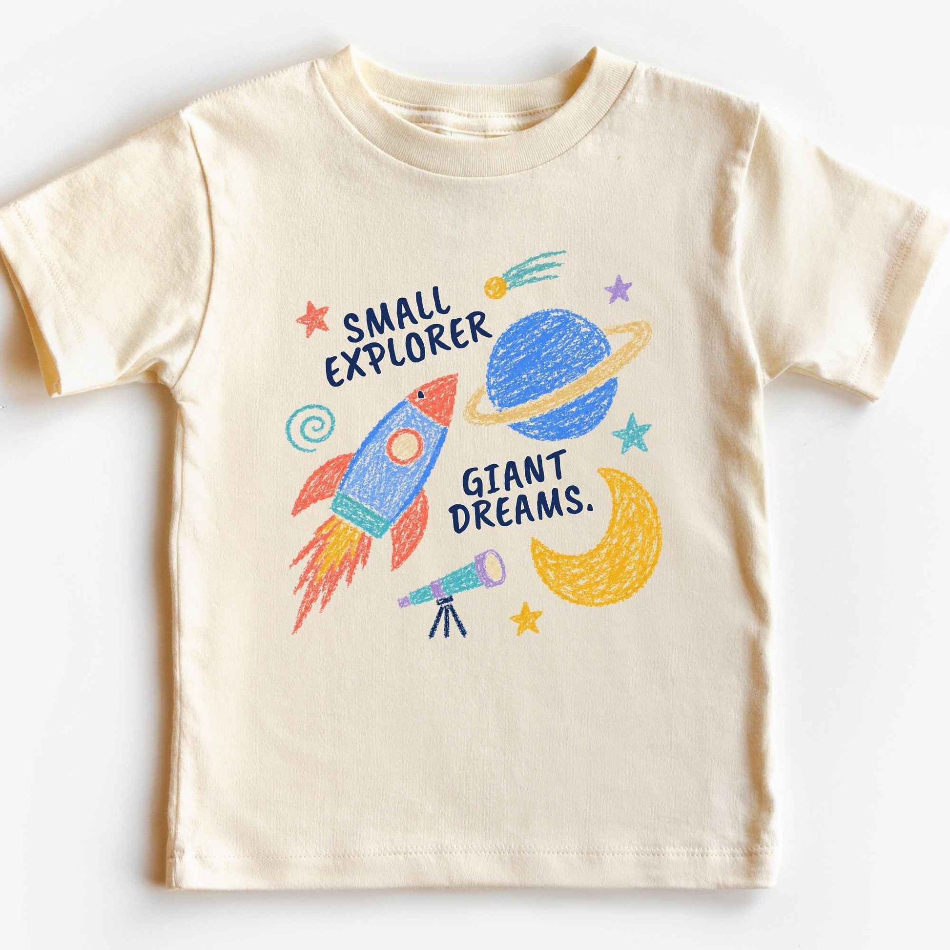 A cream-colored short-sleeve kids' t-shirt featuring a colorful hand-drawn style space graphic with a rocket, planet, moon, and stars, accompanied by the text 'SMALL EXPLORER GIANT DREAMS.'.