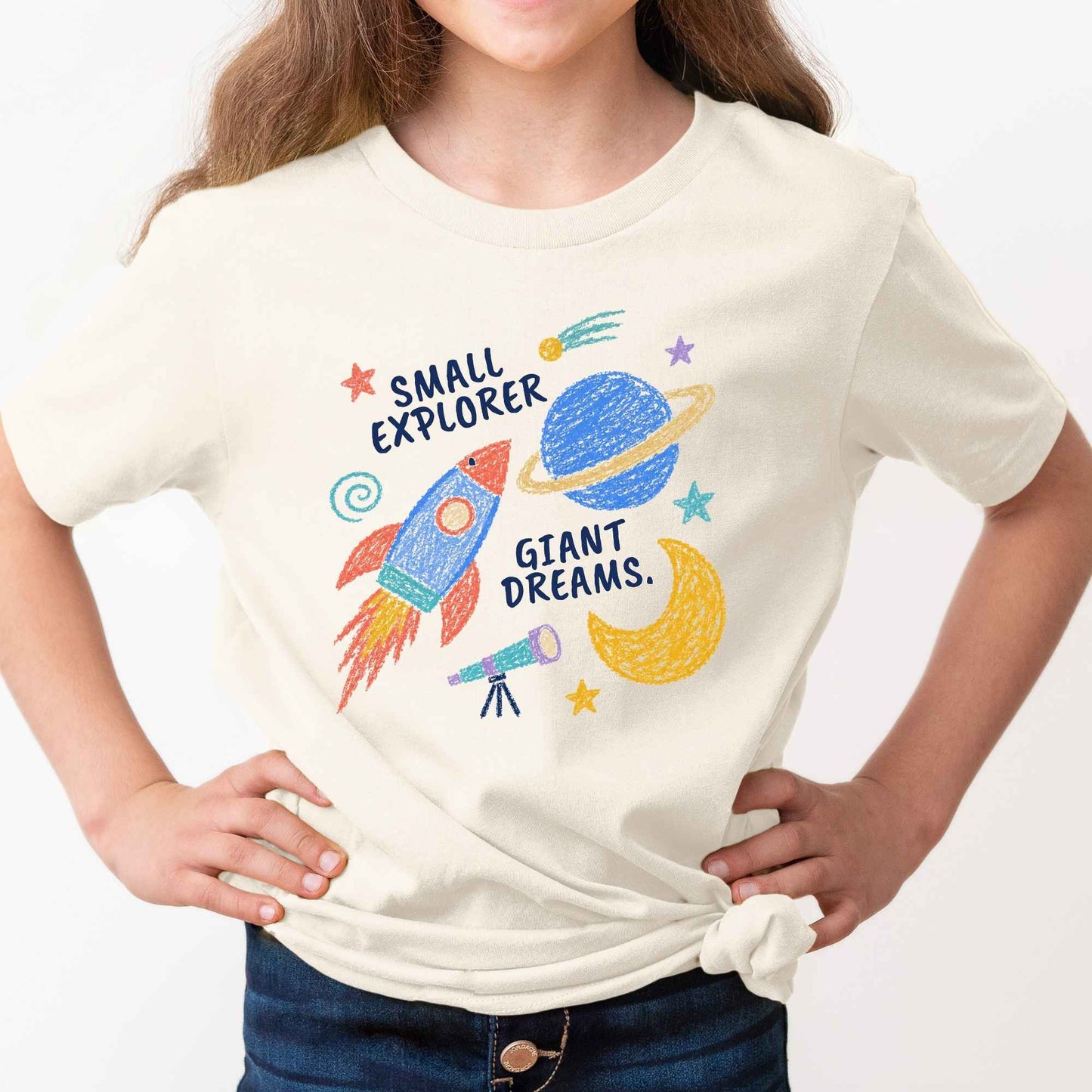 A child wearing a cream-colored t-shirt with a colorful space-themed graphic that reads 'SMALL EXPLORER GIANT DREAMS.' paired with dark blue jeans, set against a plain white background.