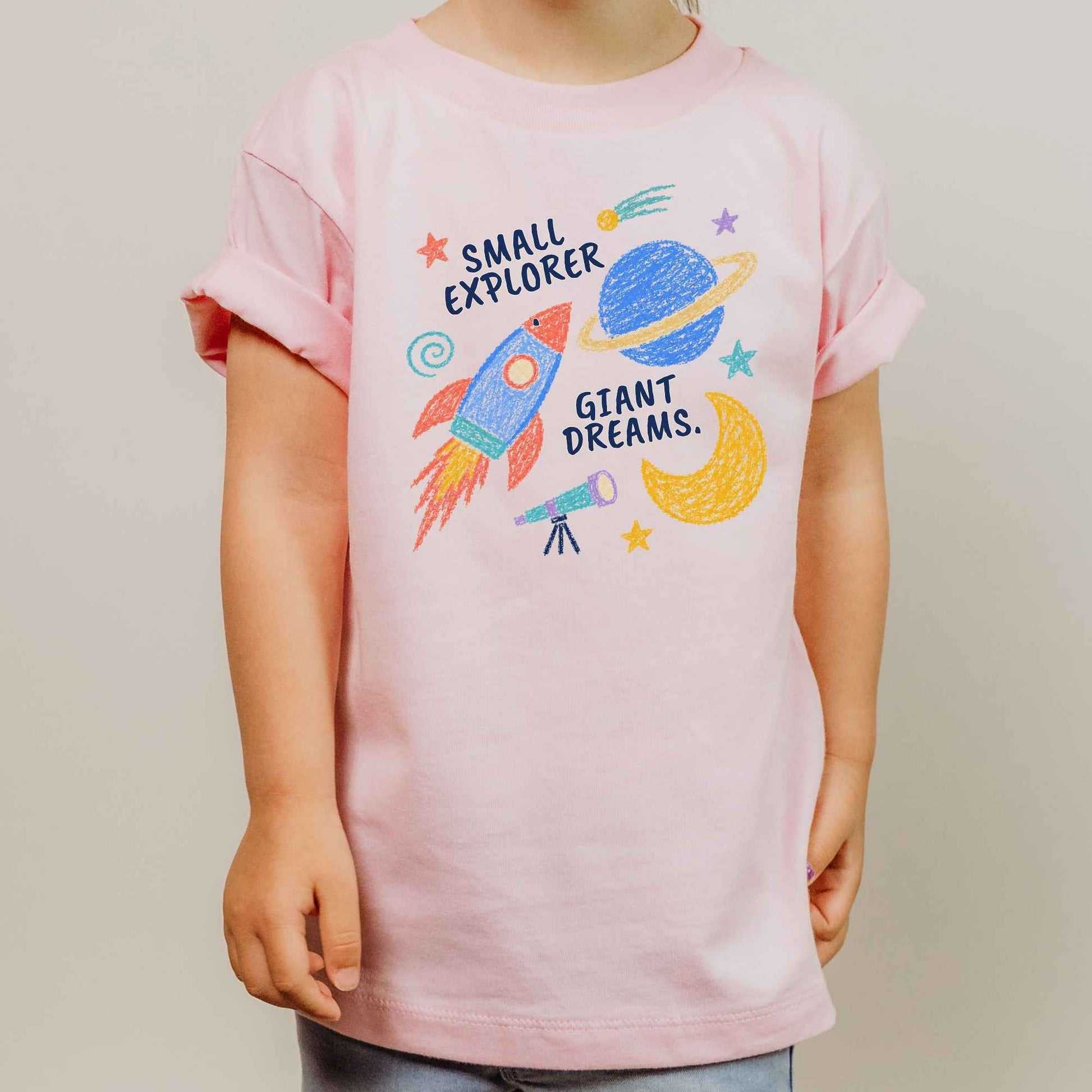 A child wearing a light pink short-sleeve t-shirt with a colorful space-themed graphic that reads 'SMALL EXPLORER GIANT DREAMS.' on a plain light gray background.