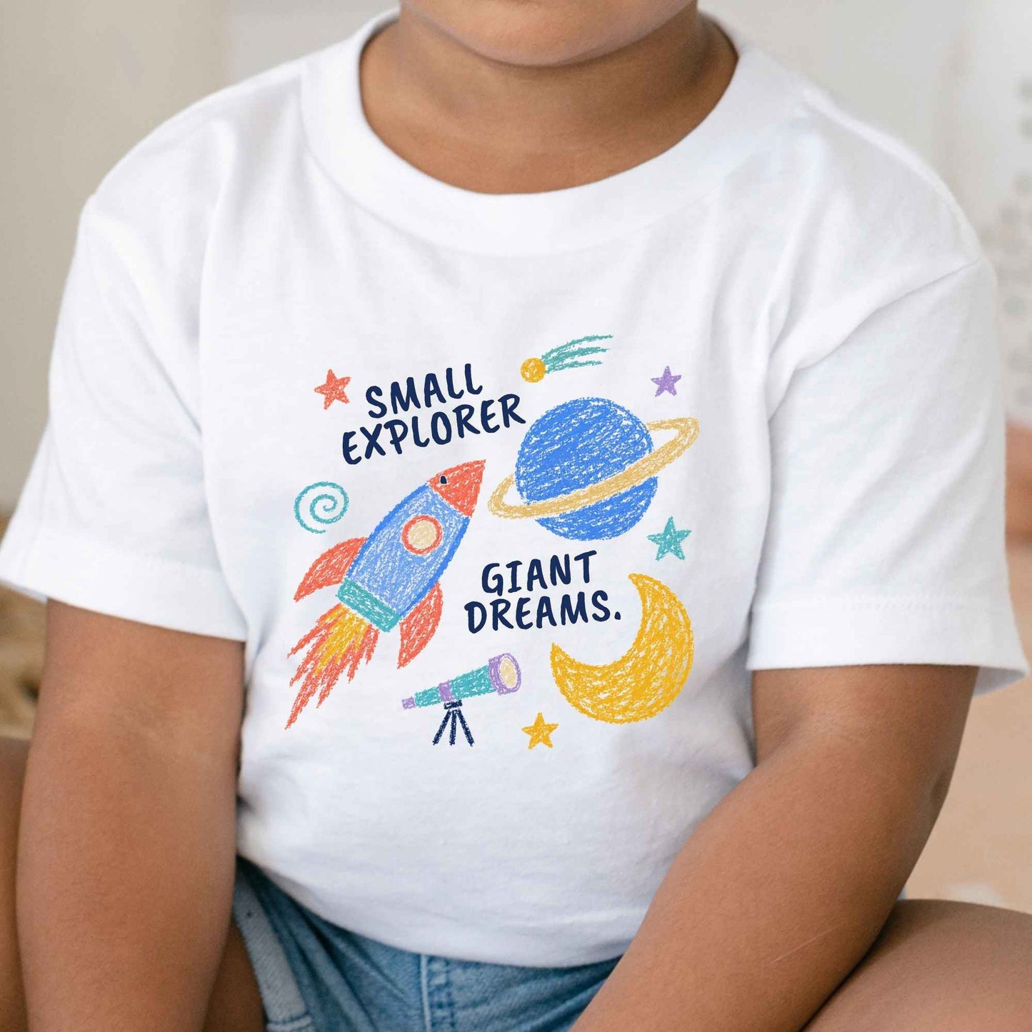 A child wearing a white short-sleeve T-shirt with a colorful space-themed graphic that reads 'SMALL EXPLORER GIANT DREAMS.' featuring a rocket, planet, moon, and stars.