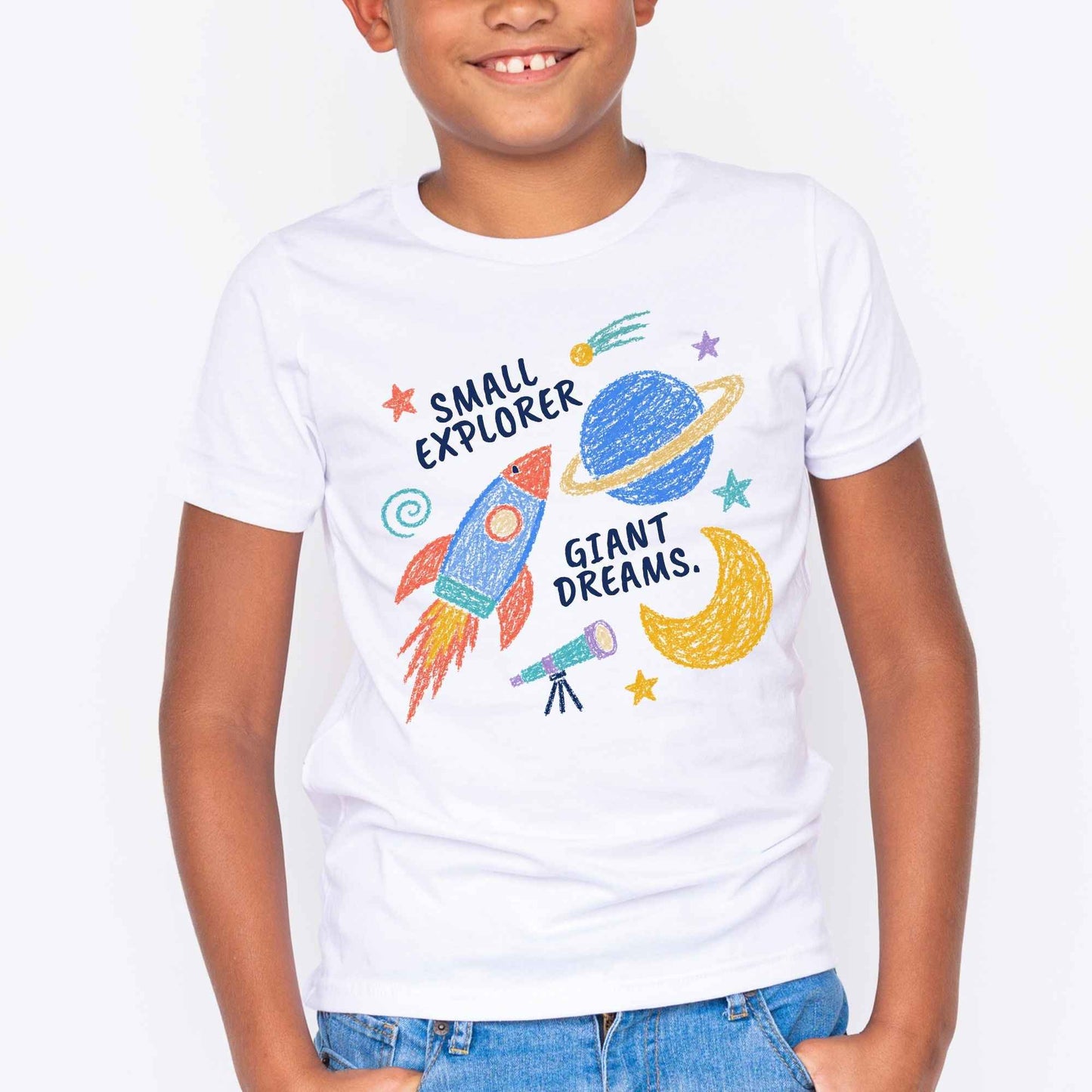 A child wearing a white short-sleeve graphic T-shirt with a colorful space-themed print that reads 'SMALL EXPLORER GIANT DREAMS.' and includes a rocket, planet, moon, and stars, paired with blue jeans against a plain white background.
