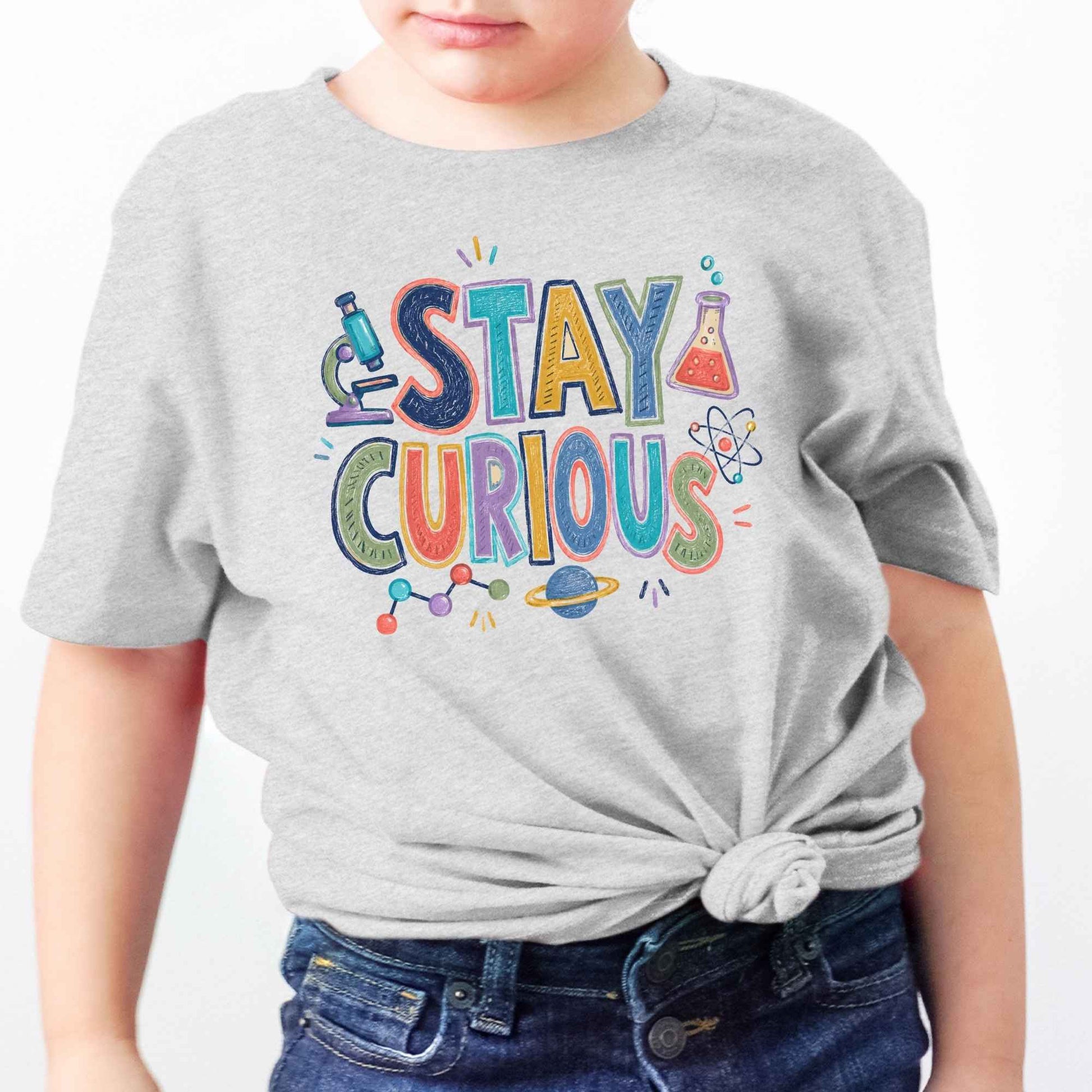 A child wearing a light gray heather t-shirt with a colorful 'STAY CURIOUS' graphic on the front, styled with a knot at the waist and paired with blue jeans against a plain white background.
