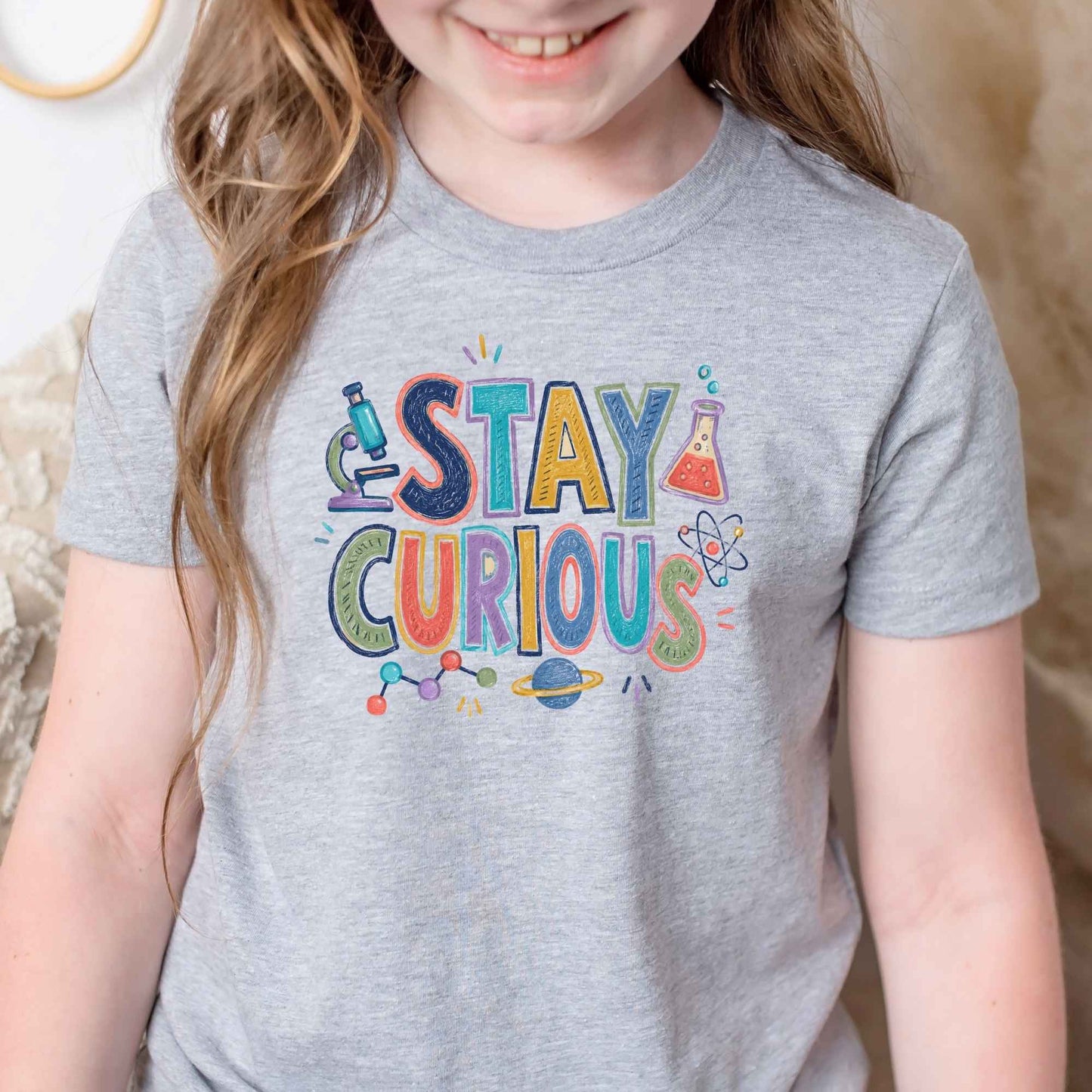 A child wearing a heather gray t-shirt with a colorful 'STAY CURIOUS' graphic printed on the chest, set against a soft indoor background.