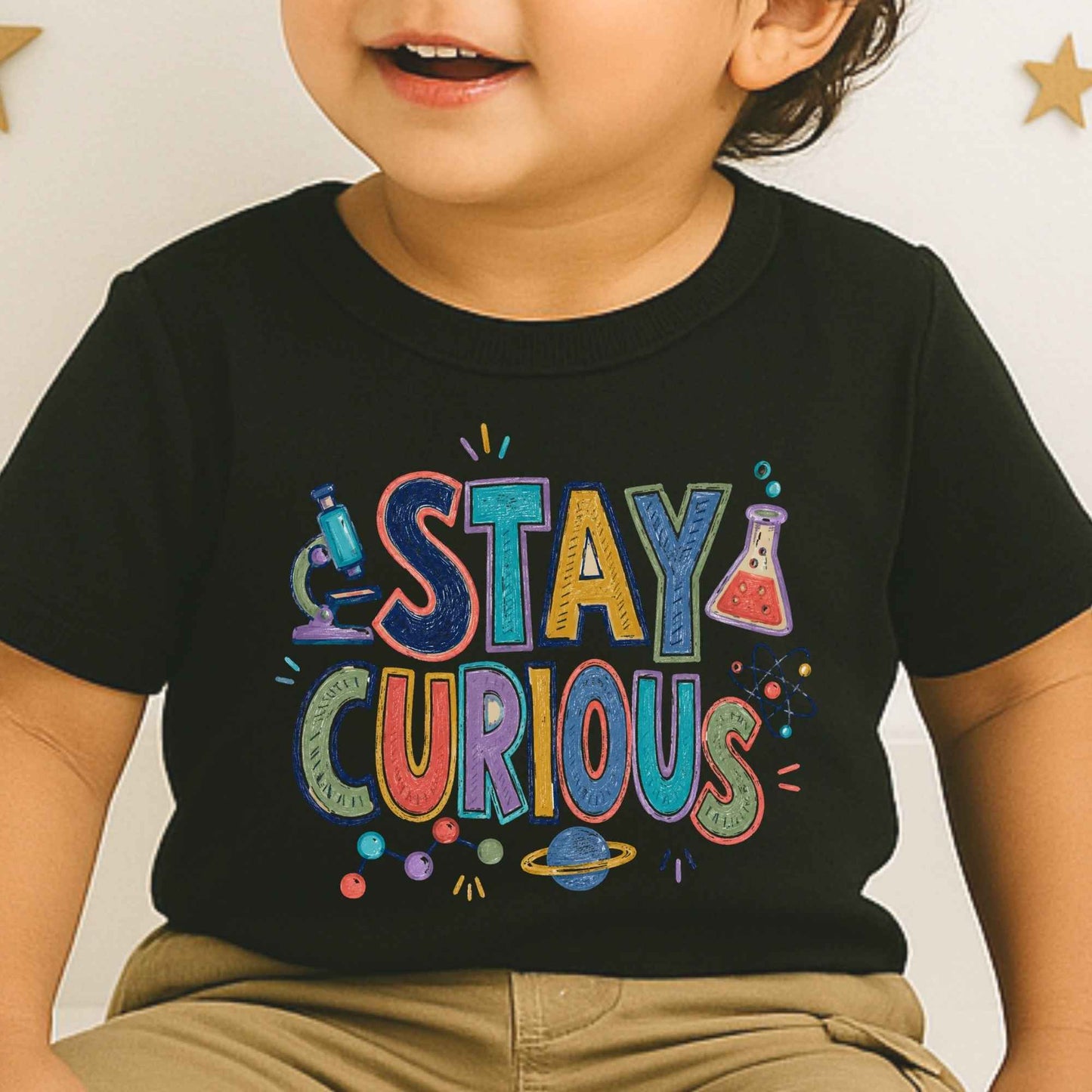 A close-up of a child wearing a black short-sleeve T-shirt with a colorful 'STAY CURIOUS' graphic on the front, paired with khaki pants against a light wall with small gold star decorations.
