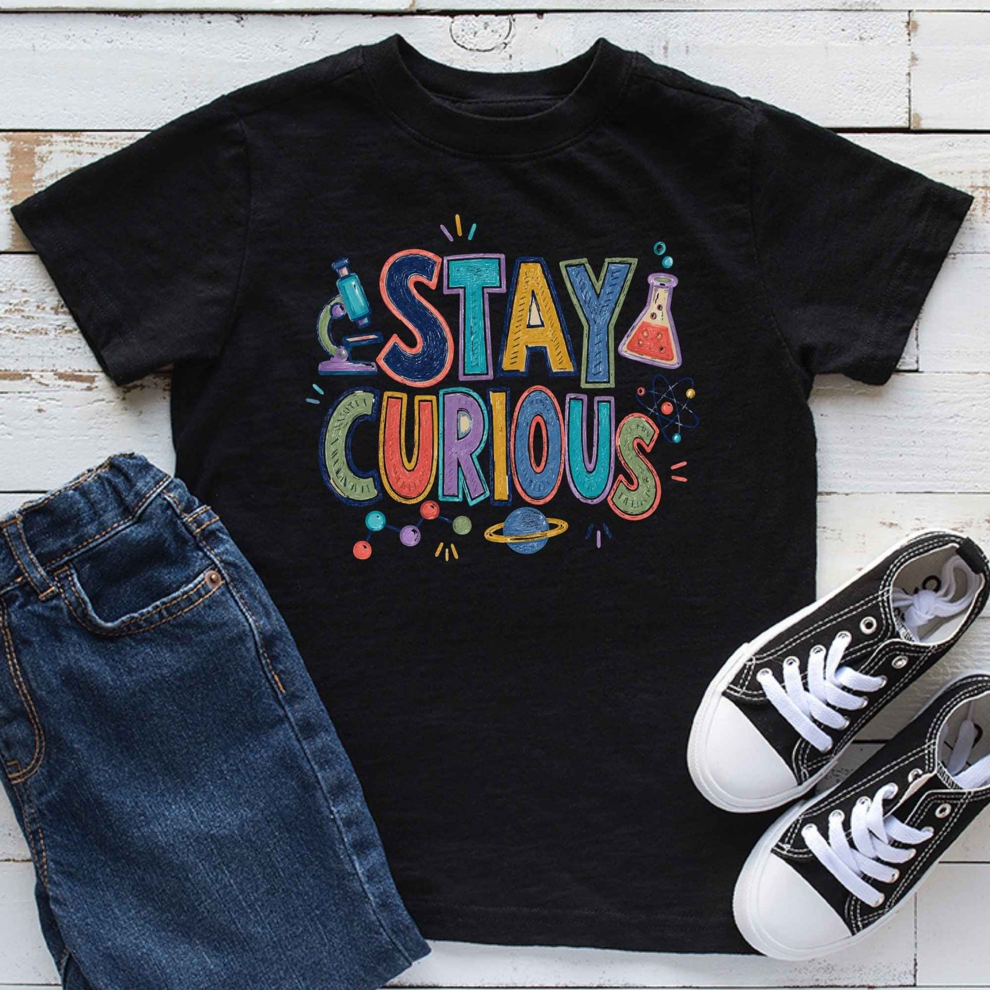 A black short-sleeve graphic T-shirt laid flat on a white wood background, styled with blue jeans and black-and-white sneakers. The shirt features a colorful retro-style 'STAY CURIOUS' print with science-themed illustrations including a microscope, beaker, and planet.