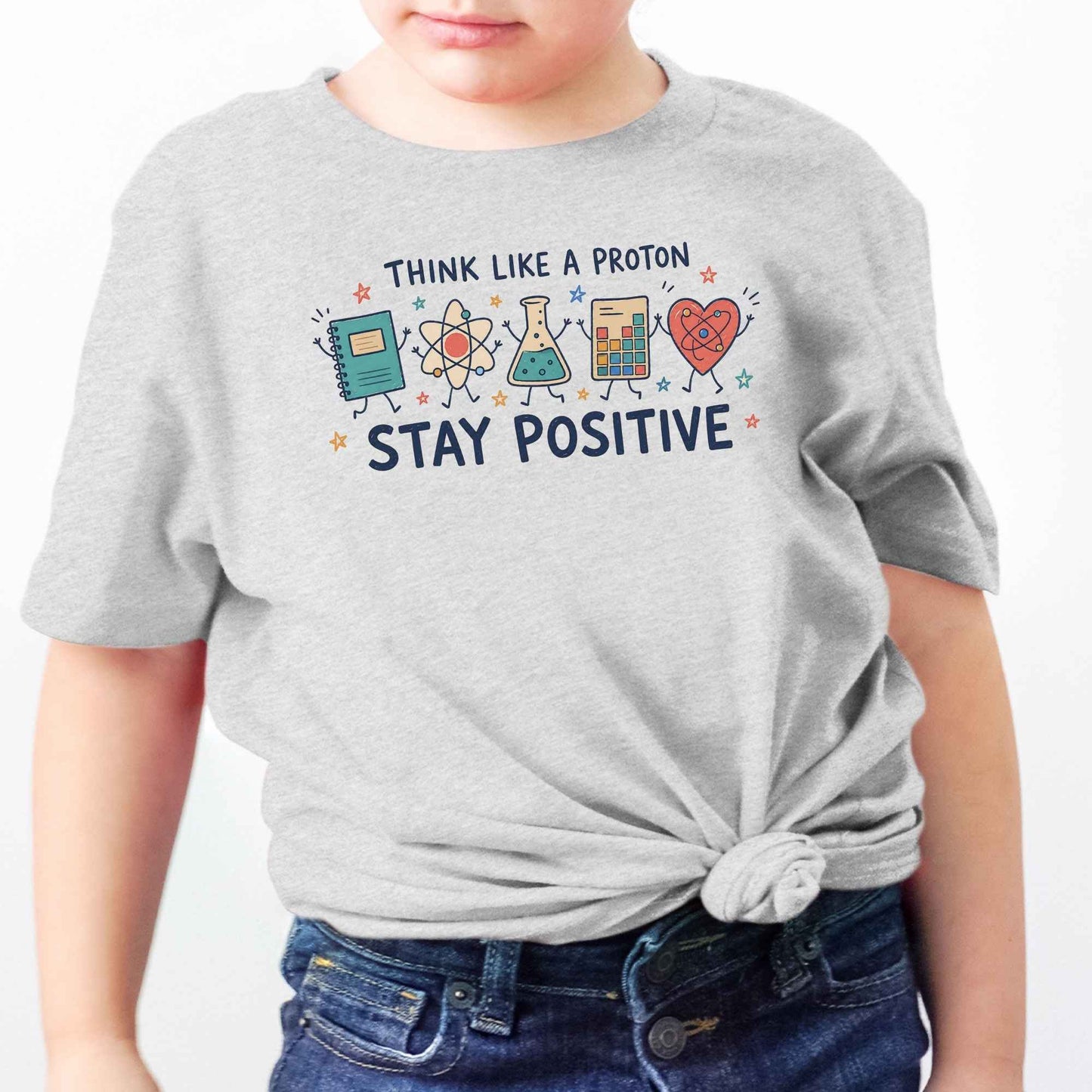 A child wearing a light heather gray t-shirt with a colorful graphic that reads 'THINK LIKE A PROTON STAY POSITIVE' with cartoon science icons, paired with blue jeans against a plain white background.