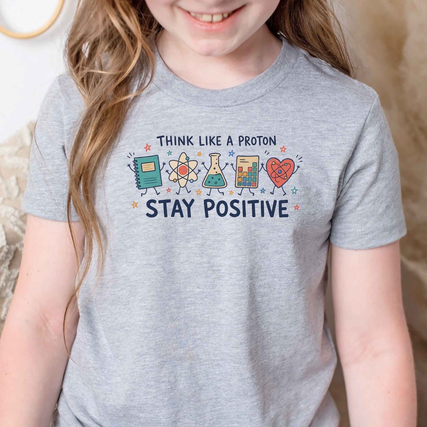 A child wearing a heather gray graphic T-shirt with the text 'THINK LIKE A PROTON STAY POSITIVE' and colorful cartoon science-themed icons on the chest, photographed indoors against a soft neutral background.