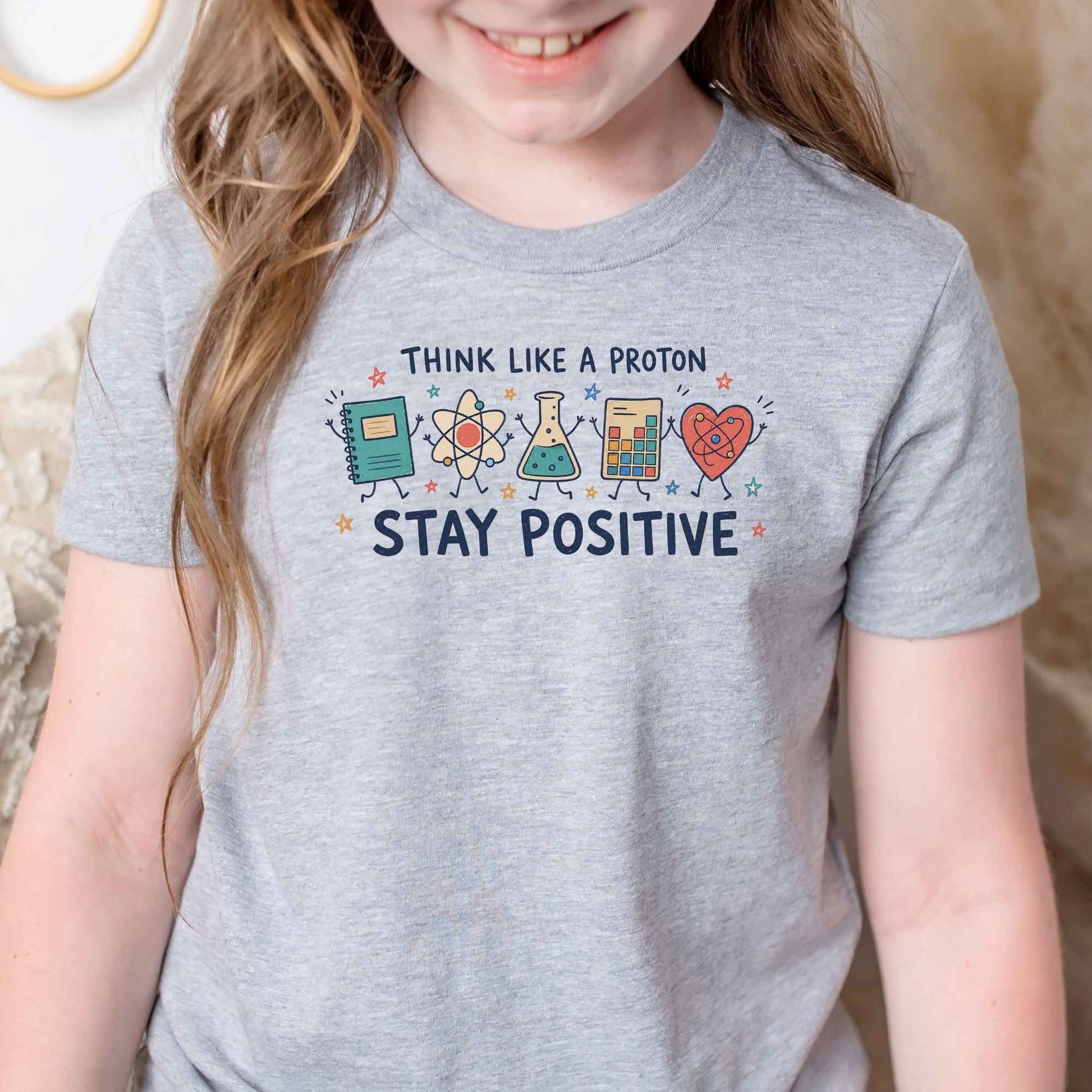 A child wearing a heather gray graphic T-shirt with the text 'THINK LIKE A PROTON STAY POSITIVE' and colorful cartoon science-themed icons on the chest, photographed indoors against a soft neutral background.