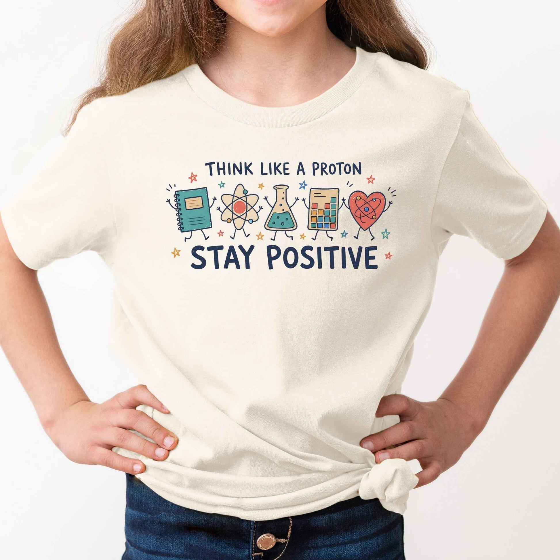 A child wearing a cream-colored t-shirt with a colorful graphic that reads 'THINK LIKE A PROTON STAY POSITIVE' accompanied by cartoon science illustrations, paired with dark blue jeans against a plain white background.