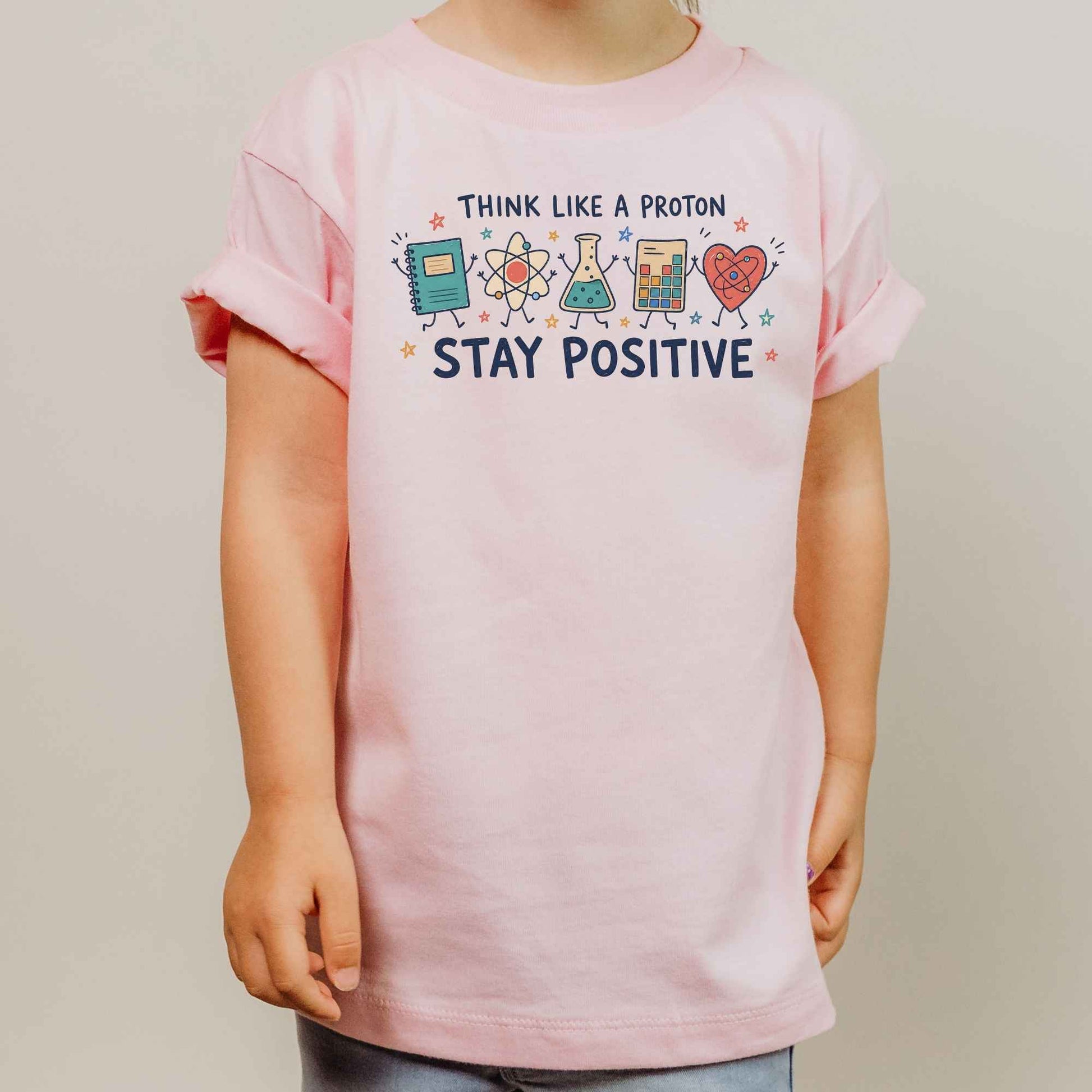 A child wearing a light pink t-shirt with a colorful graphic that reads 'THINK LIKE A PROTON STAY POSITIVE' and cartoon science symbols, photographed against a plain light gray background.