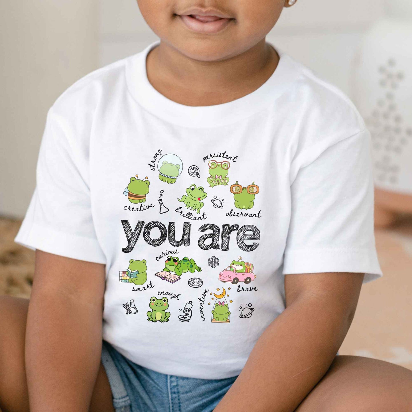 A child wearing a white short-sleeve t-shirt with a colorful frog-themed graphic that reads 'you are' in the center, paired with denim shorts in a softly lit indoor setting.