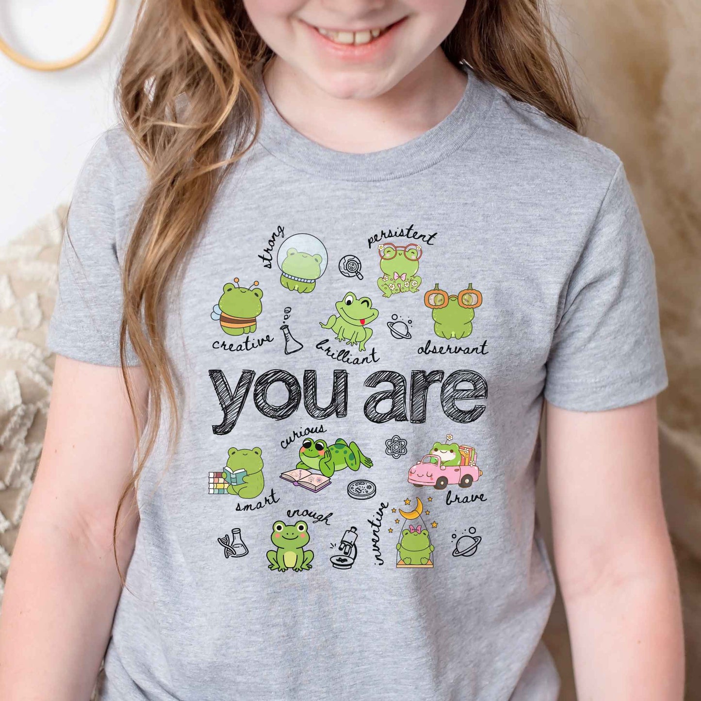 A child wearing a heather gray graphic T-shirt with cute frog illustrations and the words 'you are' printed across the front, photographed indoors against a soft neutral background.