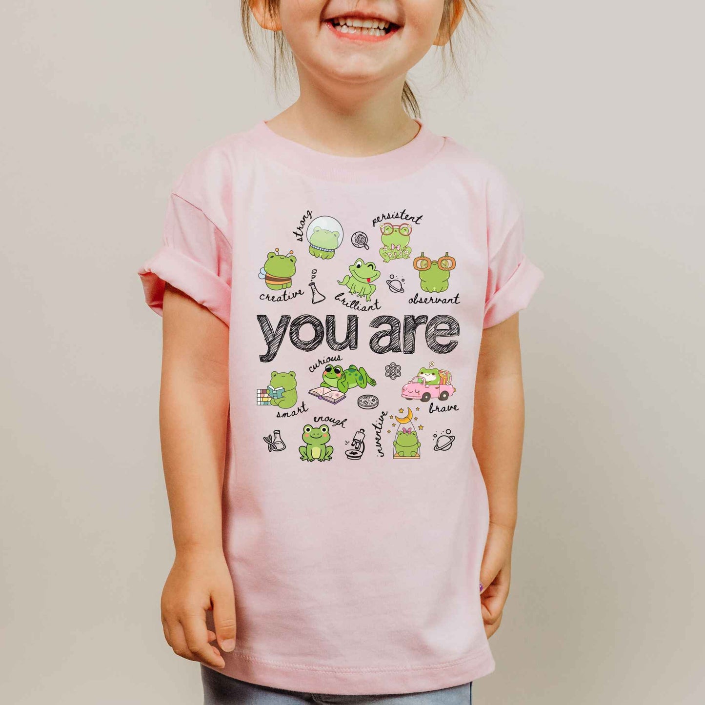 A young child wearing a light pink short-sleeve t-shirt with a cute frog-themed graphic that reads 'you are' in the center, set against a plain light gray background.
