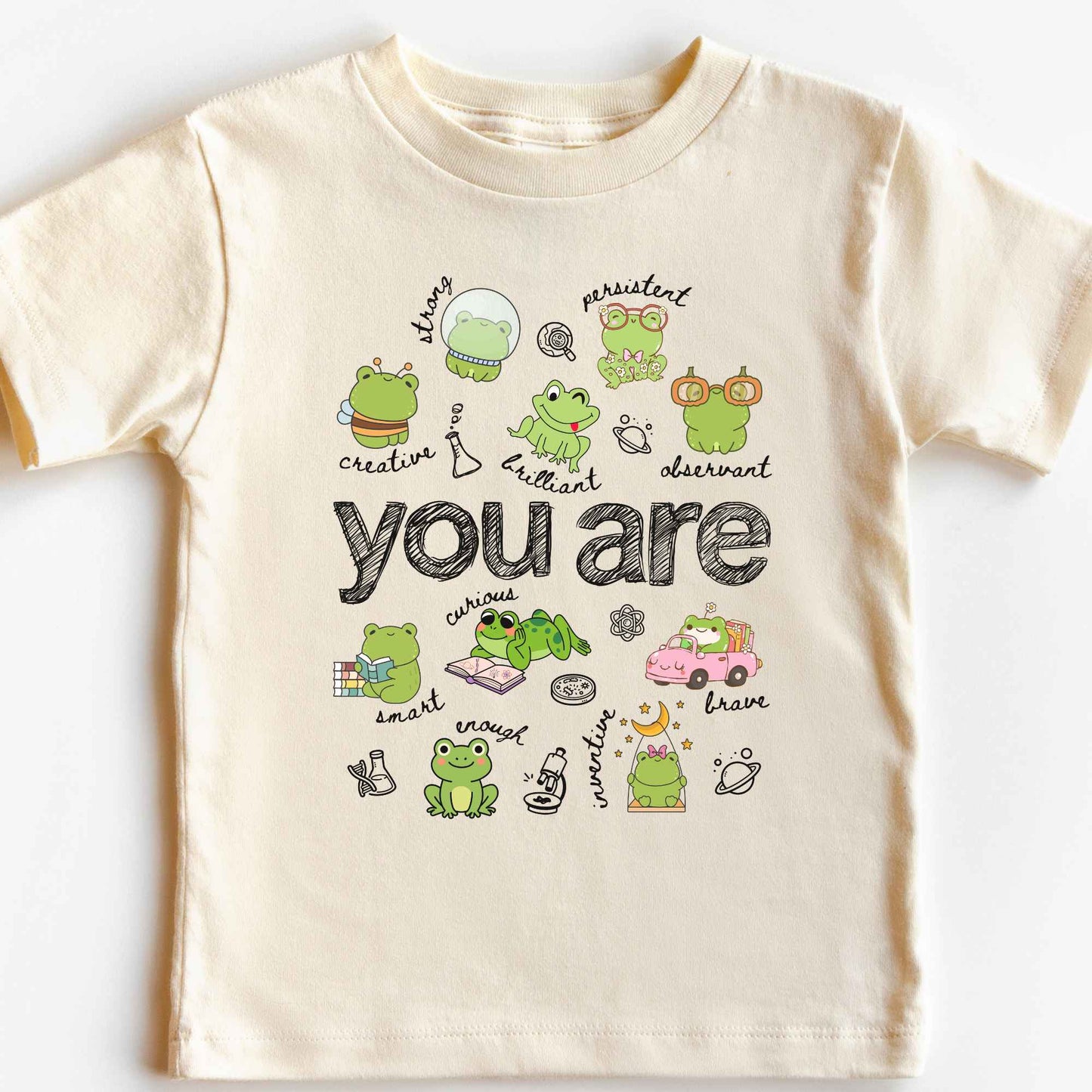 A cream-colored short-sleeve t-shirt with a cute cartoon frog graphic and the text 'you are' printed on the front, displayed flat against a white background.