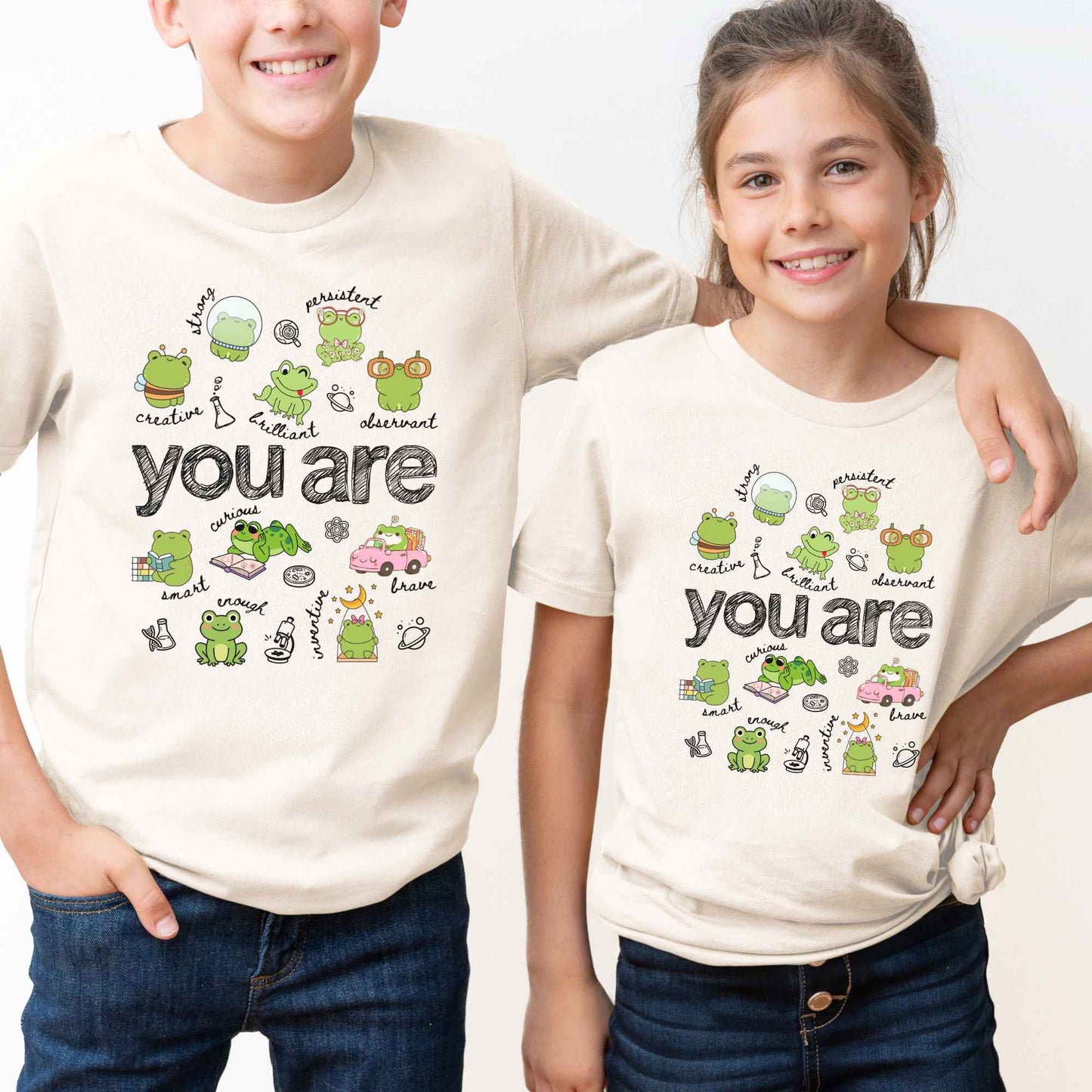 A boy and a girl wearing matching cream-colored t-shirts with a cute cartoon frog graphic and the text 'you are' on the front, set against a plain white background.