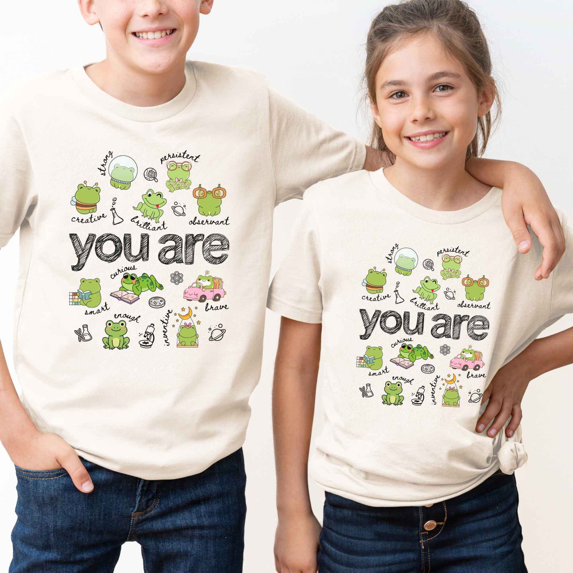 A boy and a girl wearing matching cream-colored t-shirts with a cute cartoon frog graphic and the text 'you are' on the front, set against a plain white background.