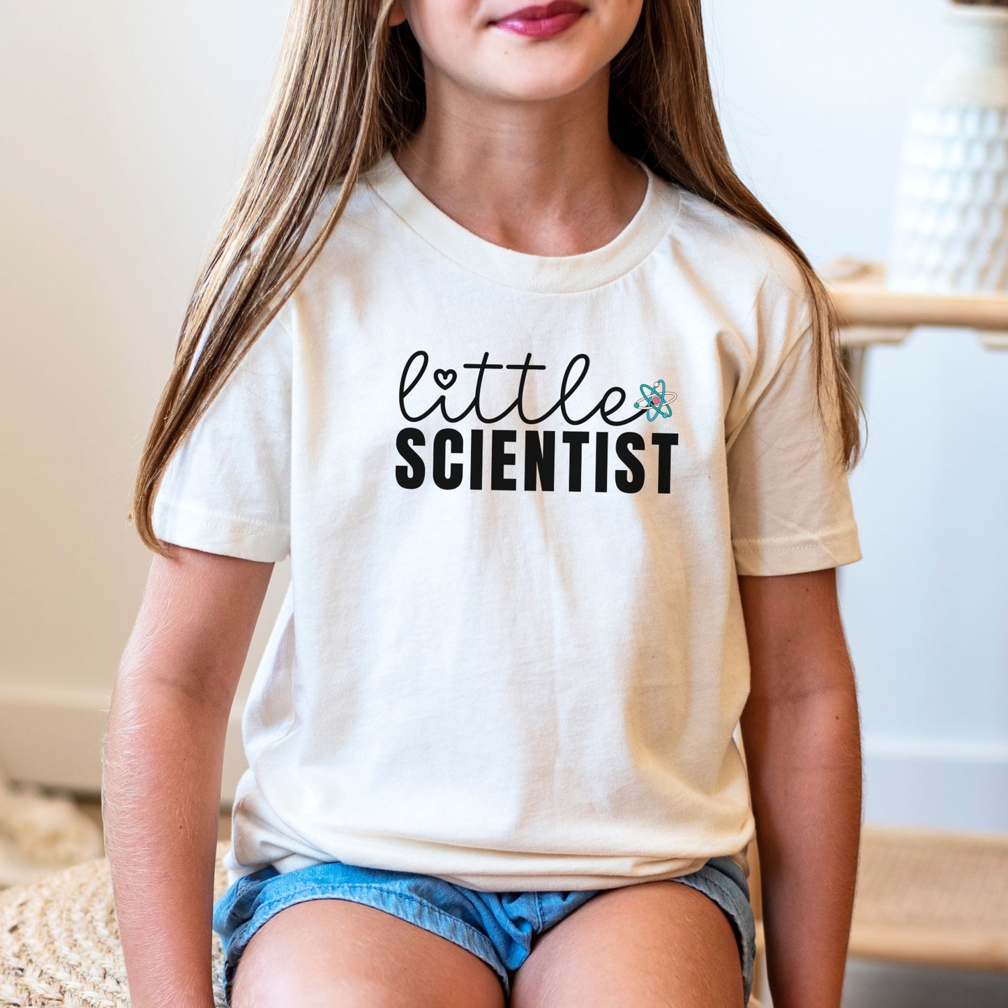 a little girl sitting on a chair wearing a t - shirt that says little scientist